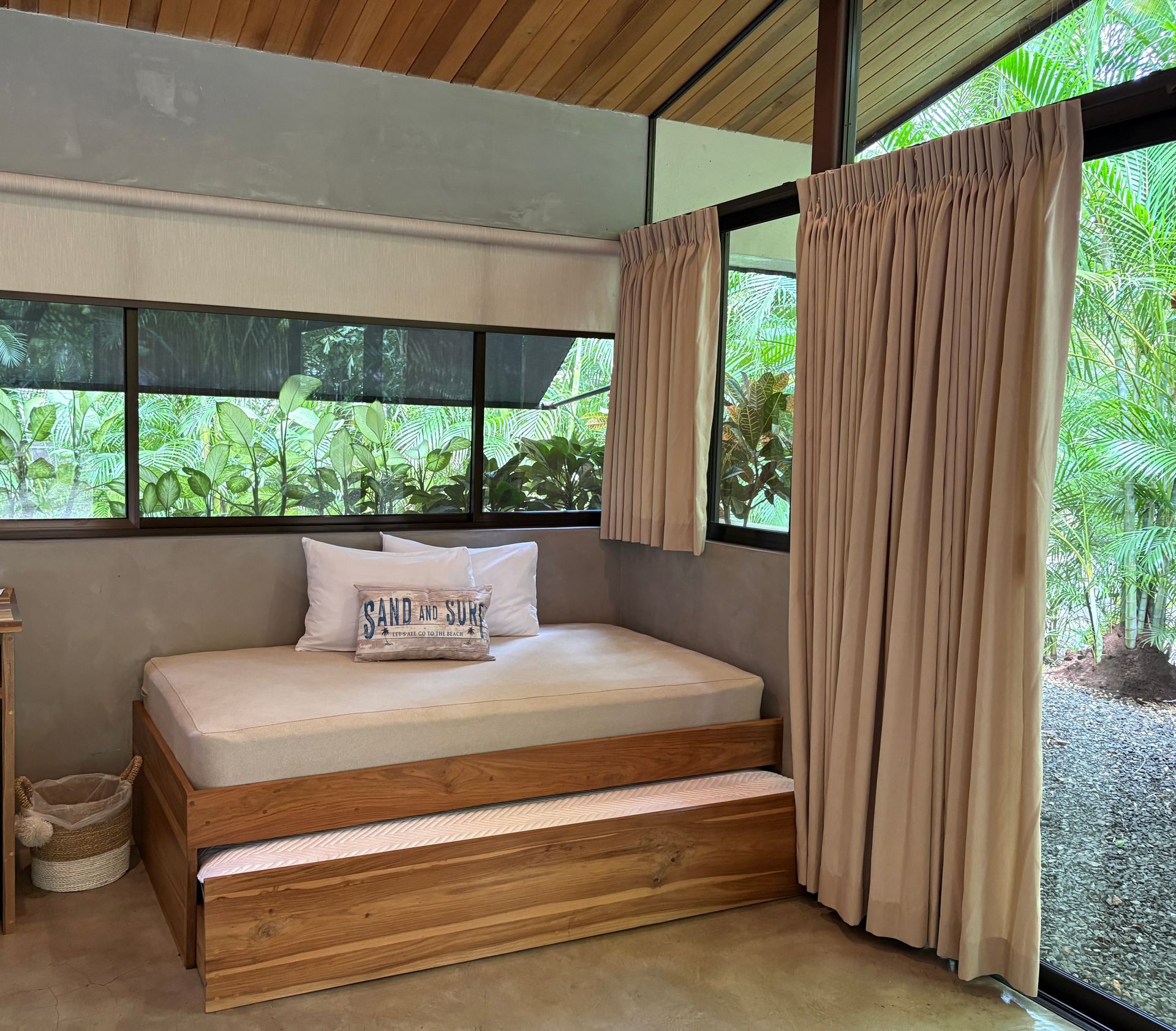 Bedroom with a trundle bed, beige curtains, and a view of greenery through the window.