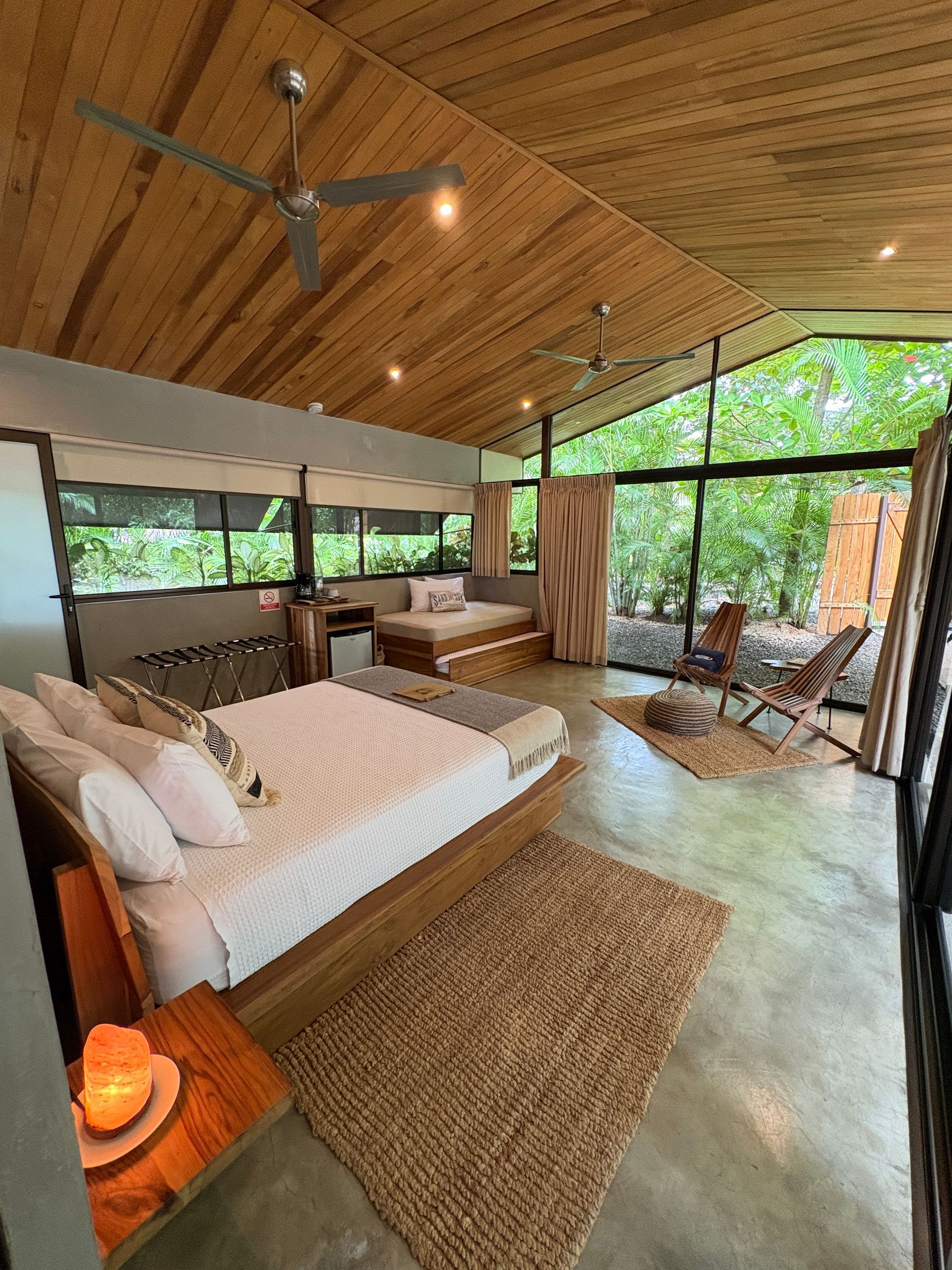 Bedroom with wood ceiling, large windows, bed, and seating area overlooking lush greenery.