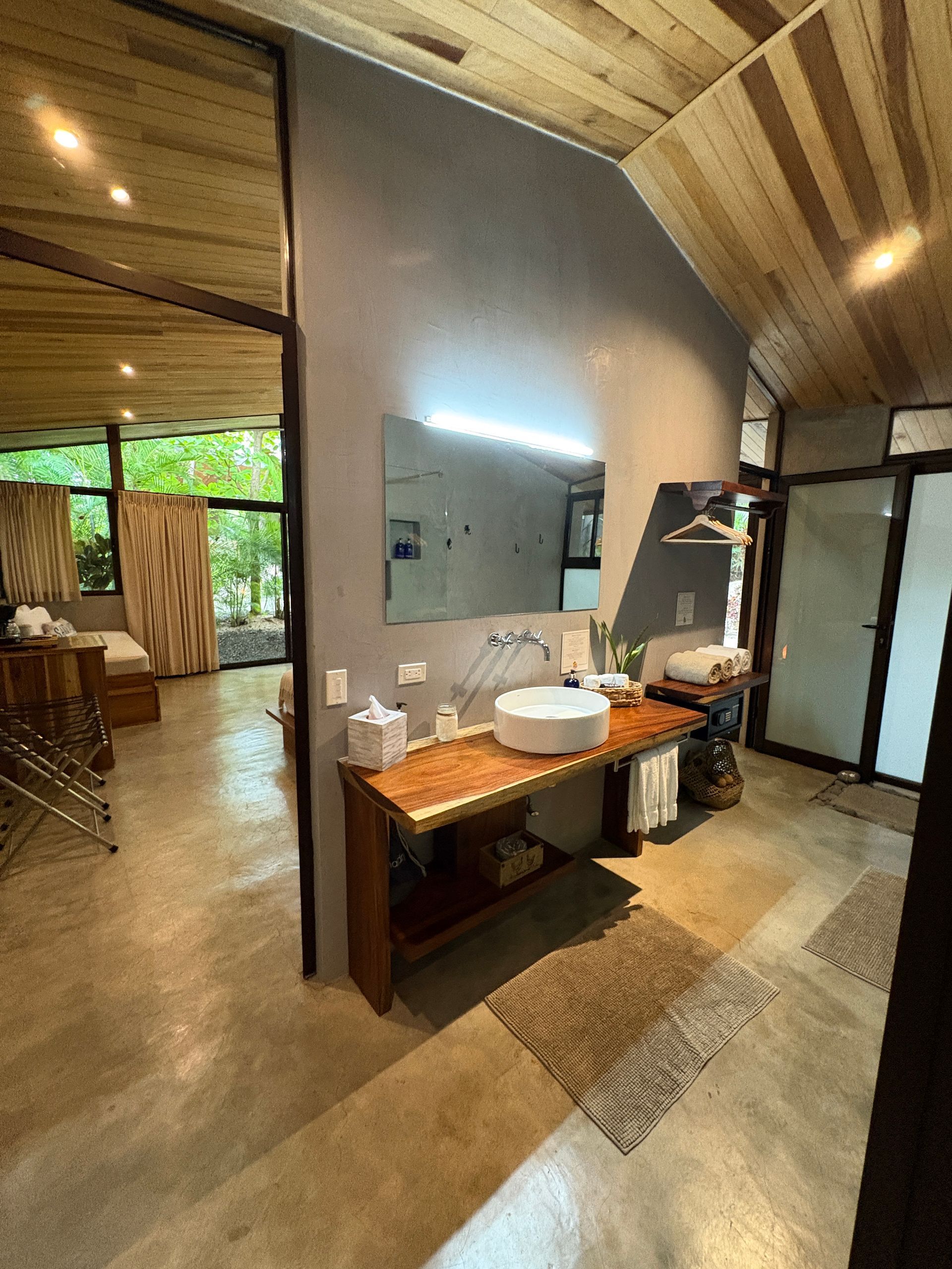 Bathroom with wooden ceiling, sink on wooden counter, open doorway to a bedroom with trees outside.