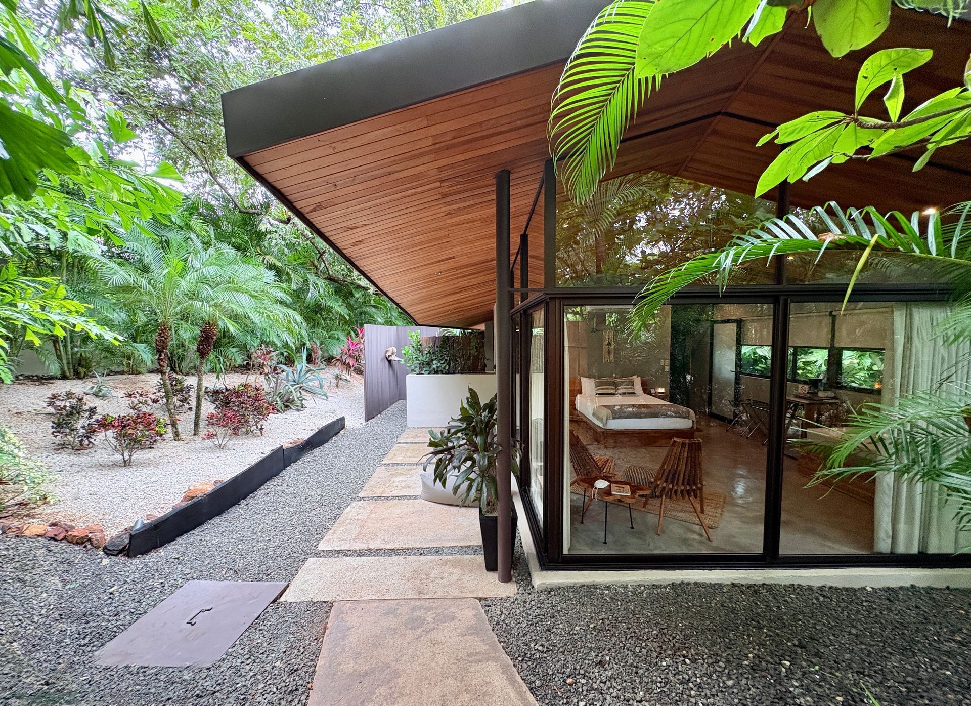 Pathway to modern glass-walled building with wood ceiling; surrounded by lush greenery and gravel.