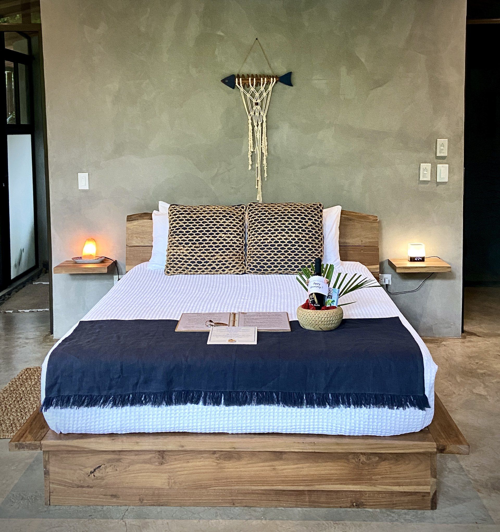 Bedroom with wooden bed, gray wall, macrame art, bedside tables with lamps, and a blanket.