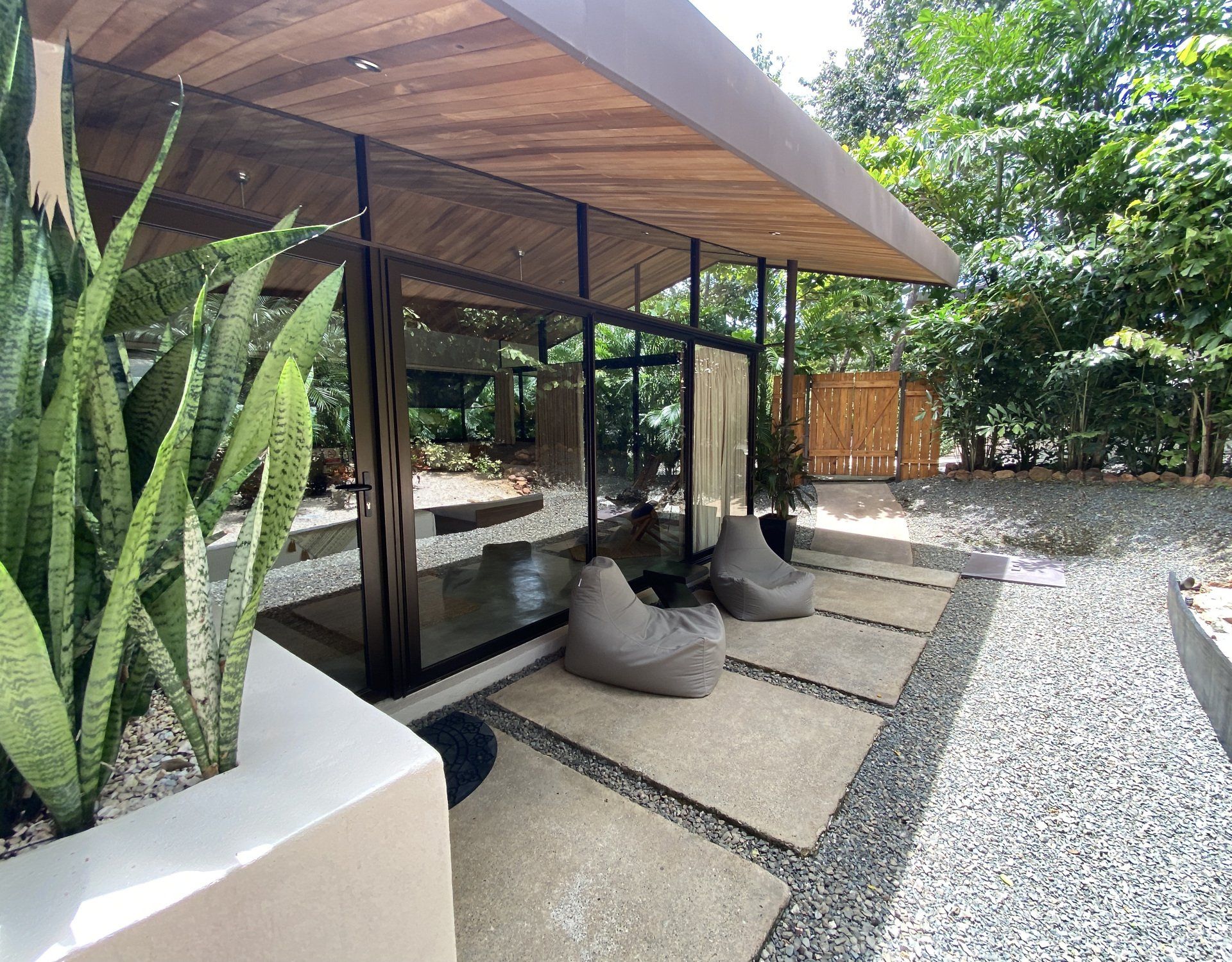 Modern outdoor lounge with glass walls, wooden ceiling, and bean bag chairs on a stone patio.