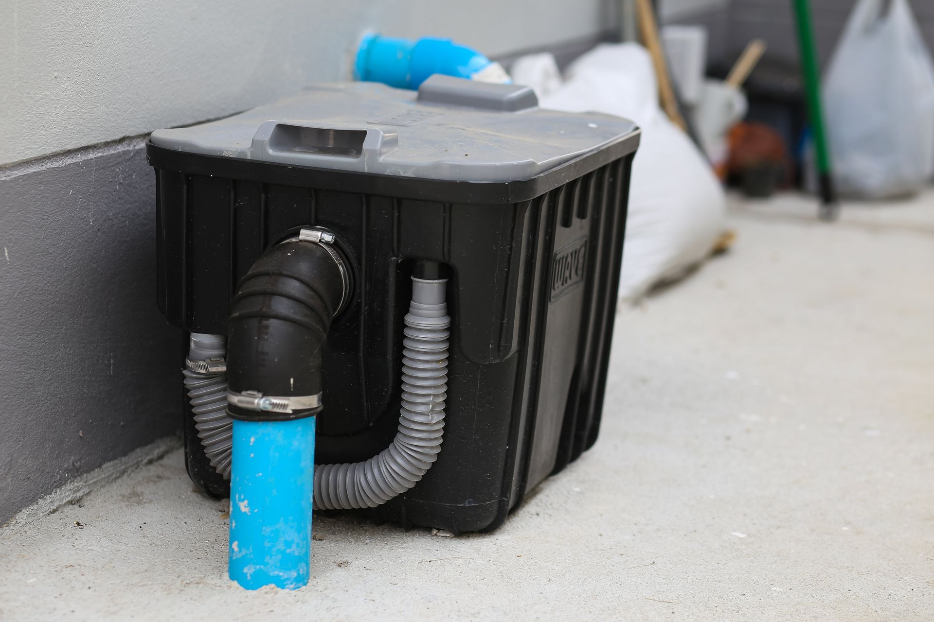 Black, plastic container with connected pipes, next to a wall on a concrete surface.