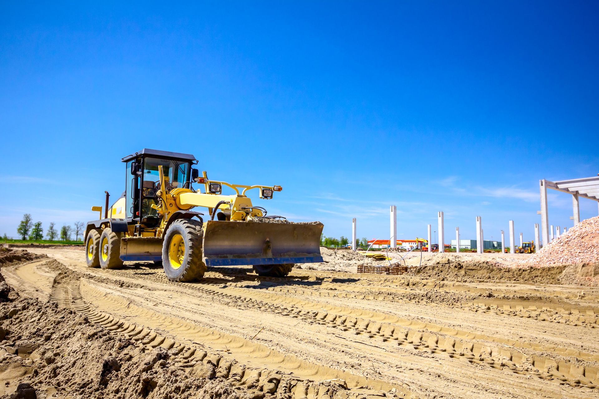 Yellow motor grader on dirt at a construction site under a blue sky.