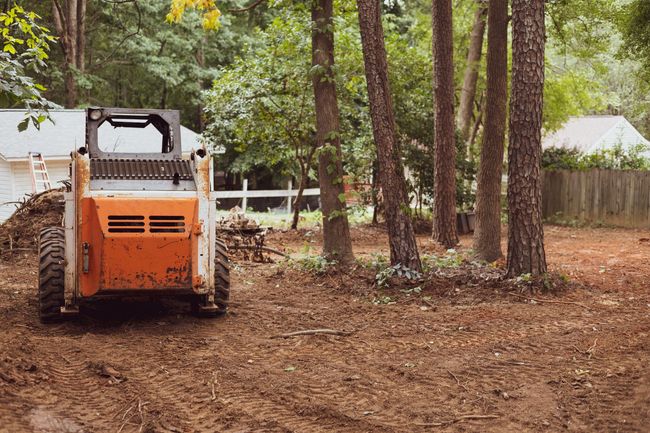 Orange skid steer clearing a dirt lot surrounded by trees, with a house visible in the background.