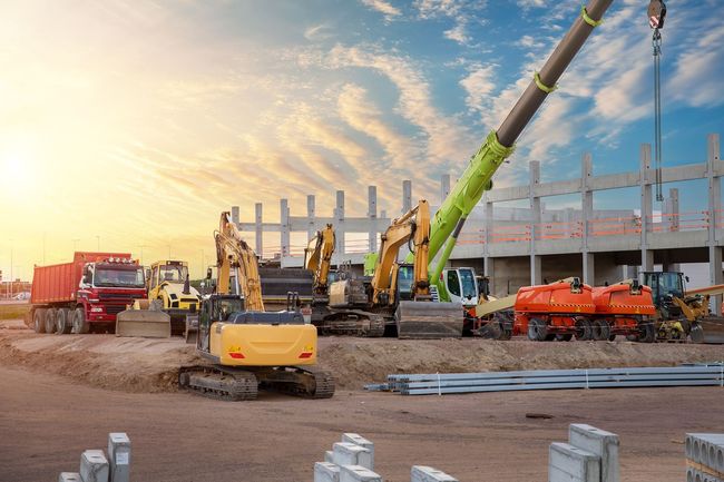 Construction site with excavators, crane, and trucks; building under construction at sunset.
