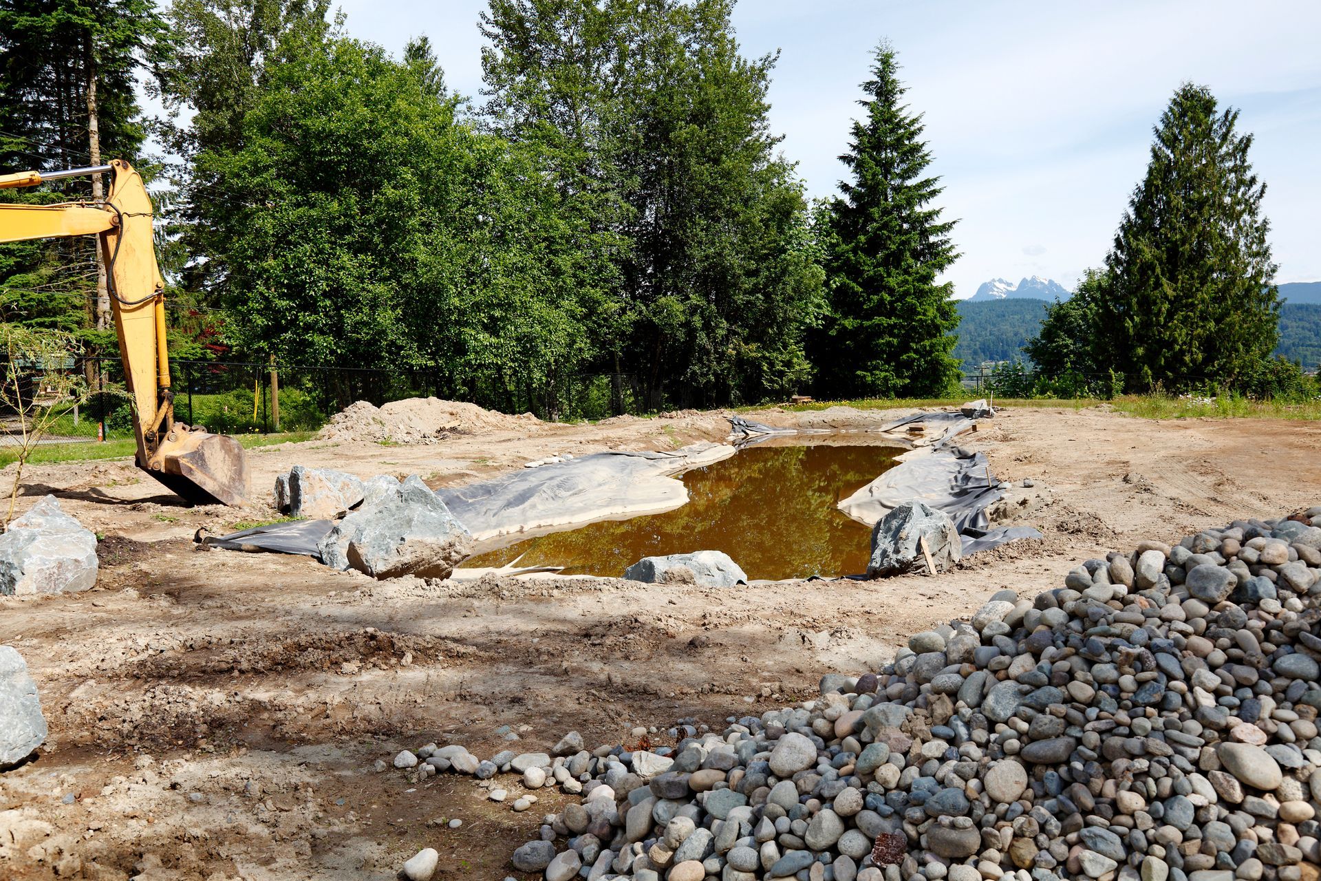 Excavated pond with stone border and murky water; excavator visible; trees and mountains in the background.