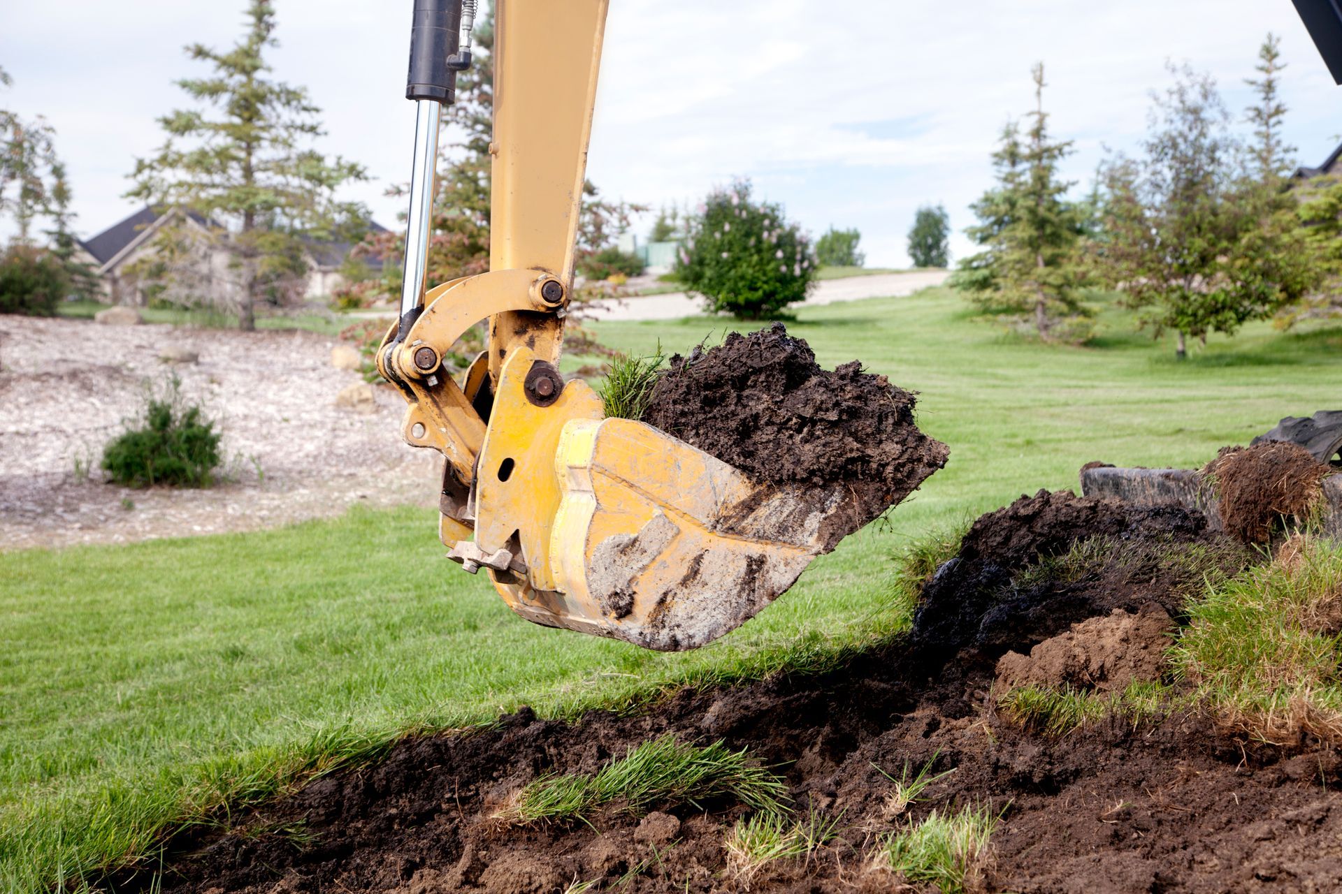 Excavator bucket filled with dark soil over a grassy area, digging a trench outdoors.