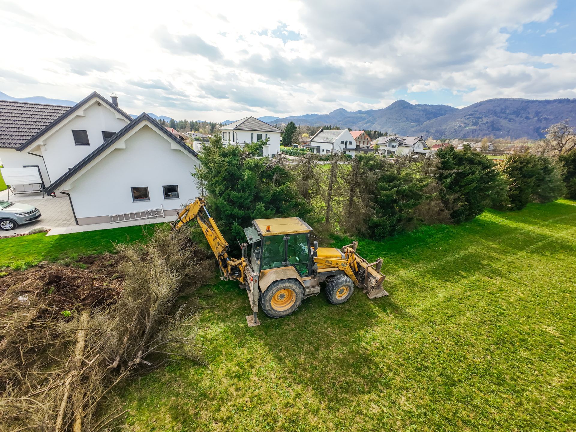 Yellow backhoe clearing brush on a green lawn near white houses, mountains in the distance.