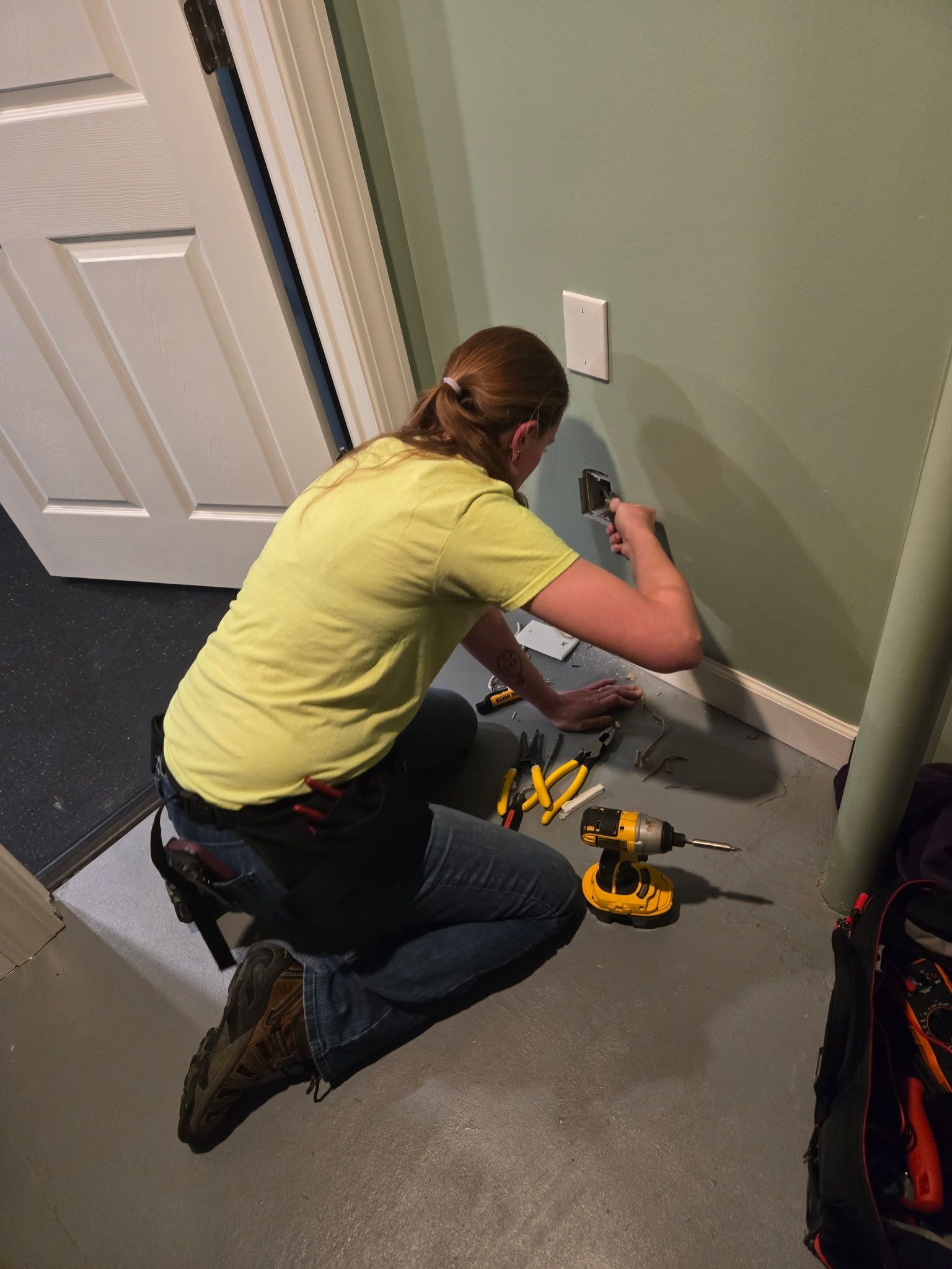 A woman is kneeling on the floor working on a wall with a drill.