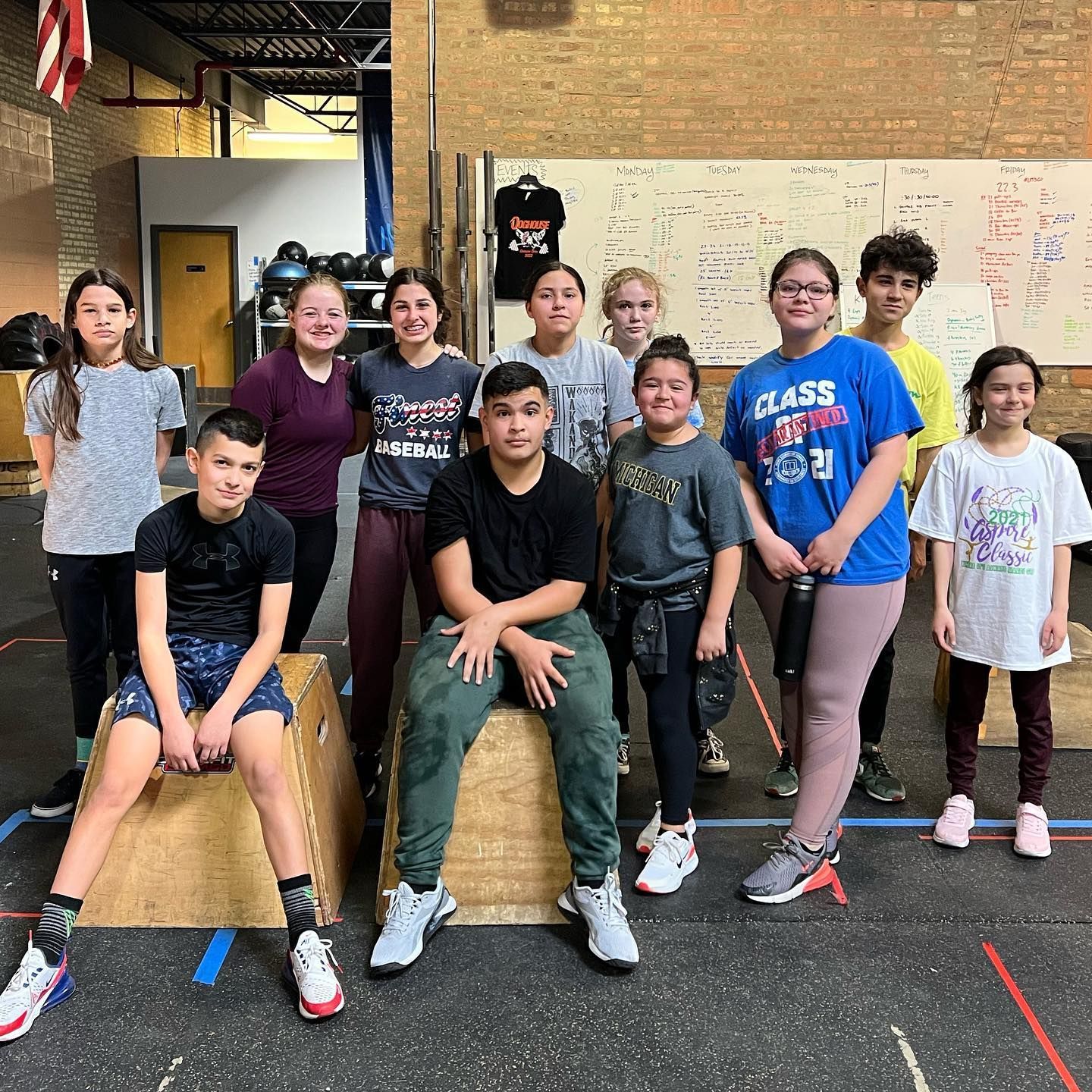 Group of kids posing in a gym, some seated on boxes. Diverse ages, wearing athletic clothes, smiling.