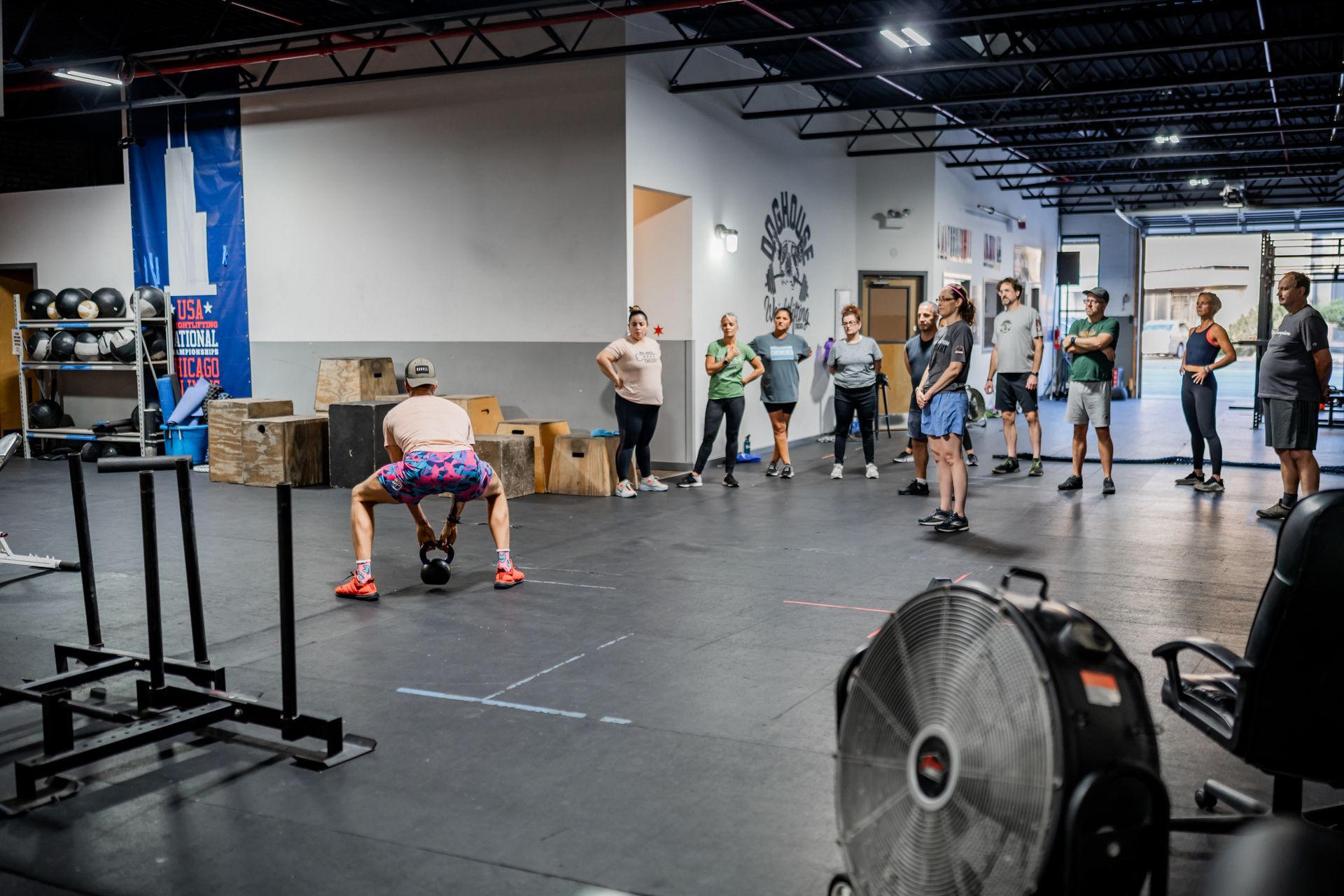 A man in colorful shorts swings a kettlebell as a group watches in a gym with exercise equipment.