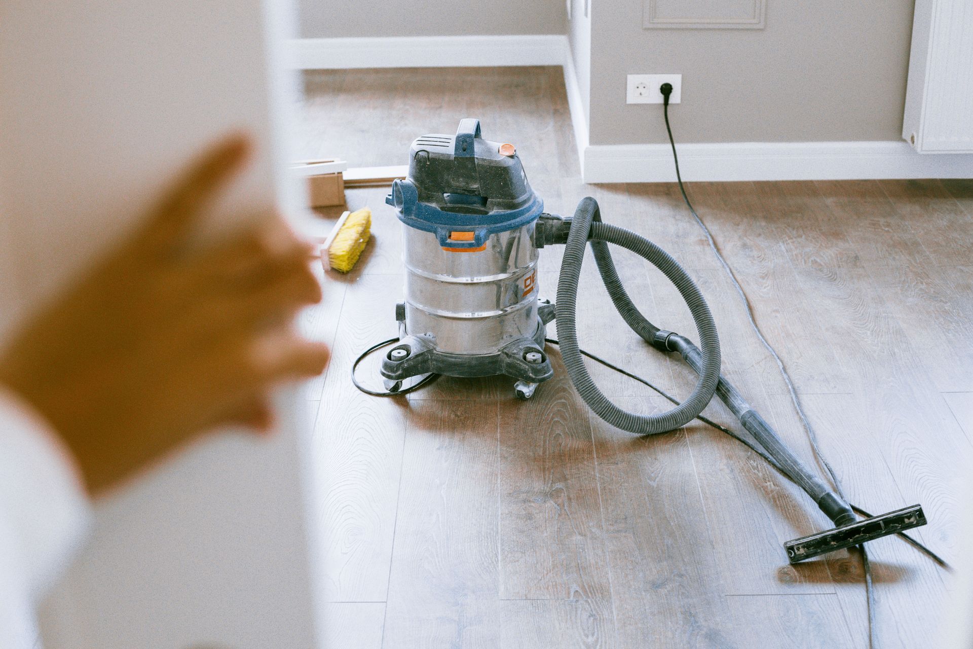 Vacuum cleaner on a hardwood floor with a hand reaching in from the left.