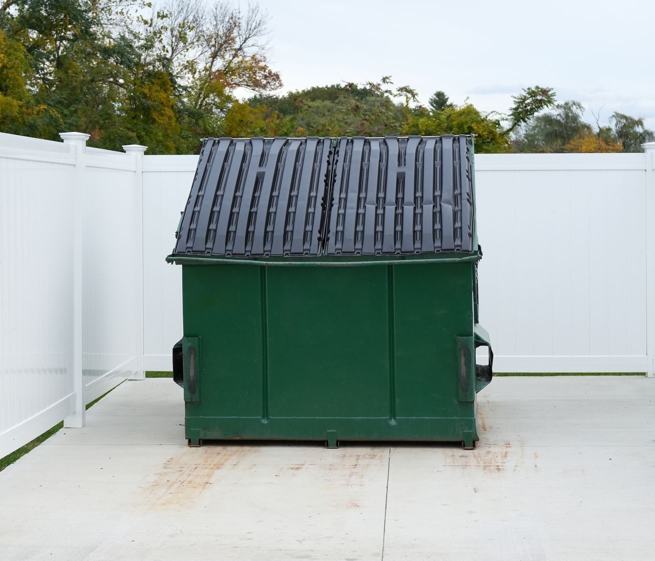 A green dumpster with a white fence in the background