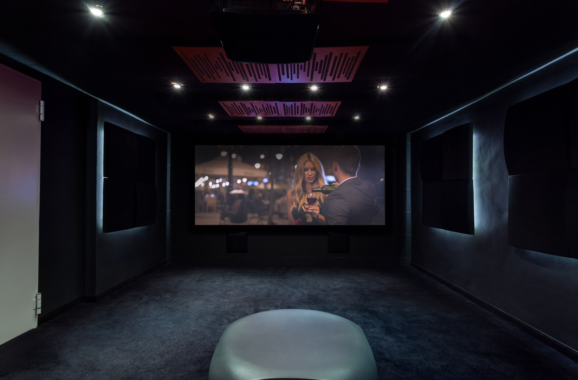 Dark home theater with large screen, sound panels, recessed lighting, black walls, and a blue carpet.