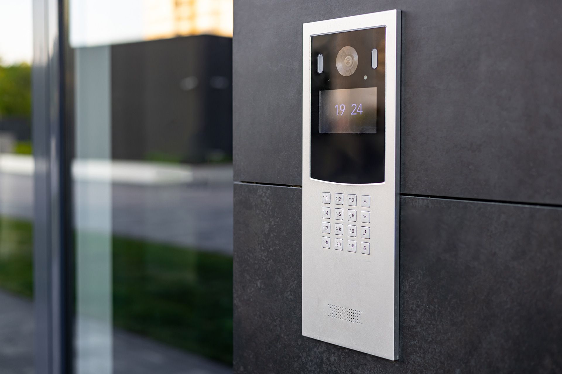 Silver intercom panel with a keypad and screen on a dark gray wall, near glass.