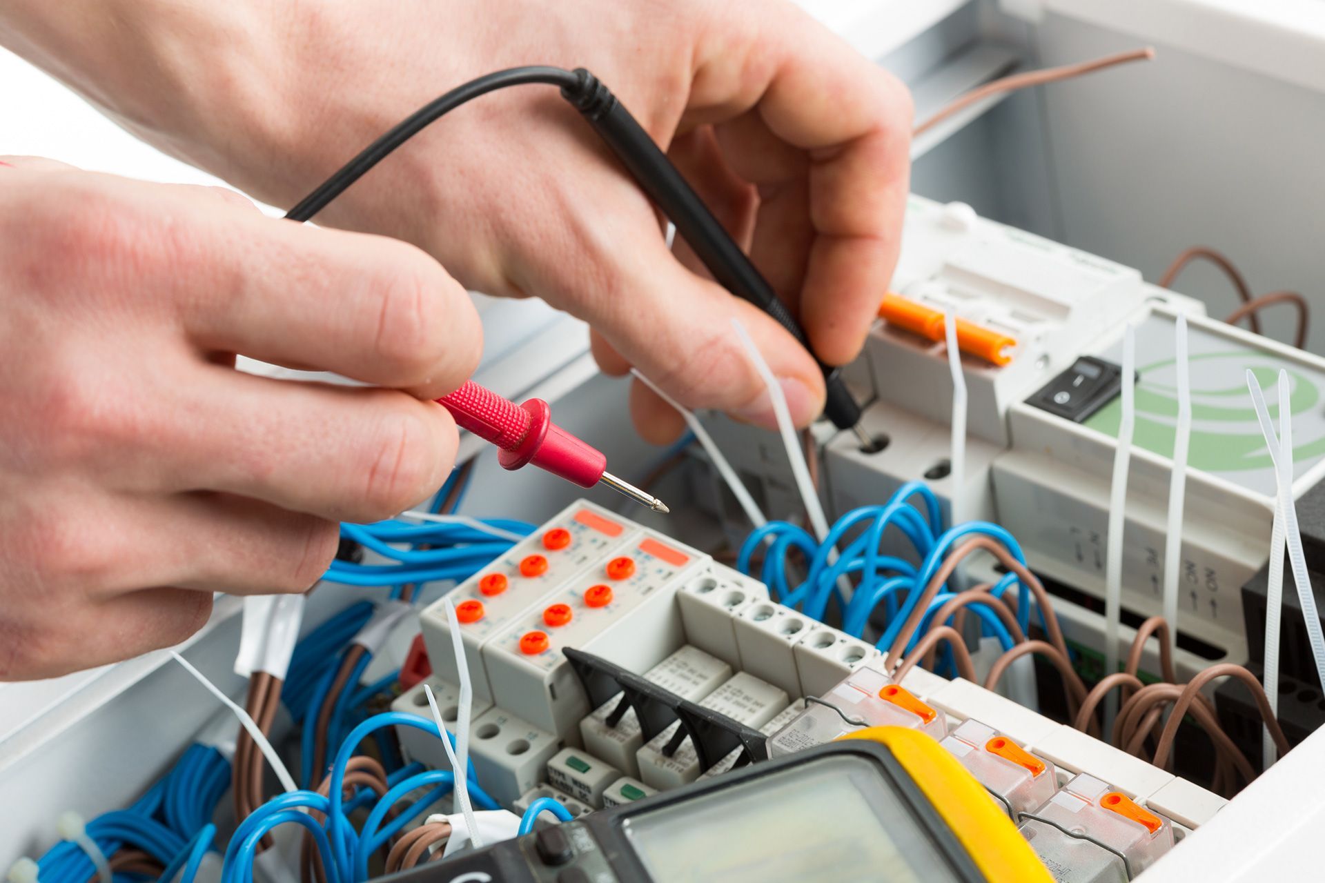 Hands of an electrician using a multimeter to test wires inside an electrical panel; blue and orange wires are visible.