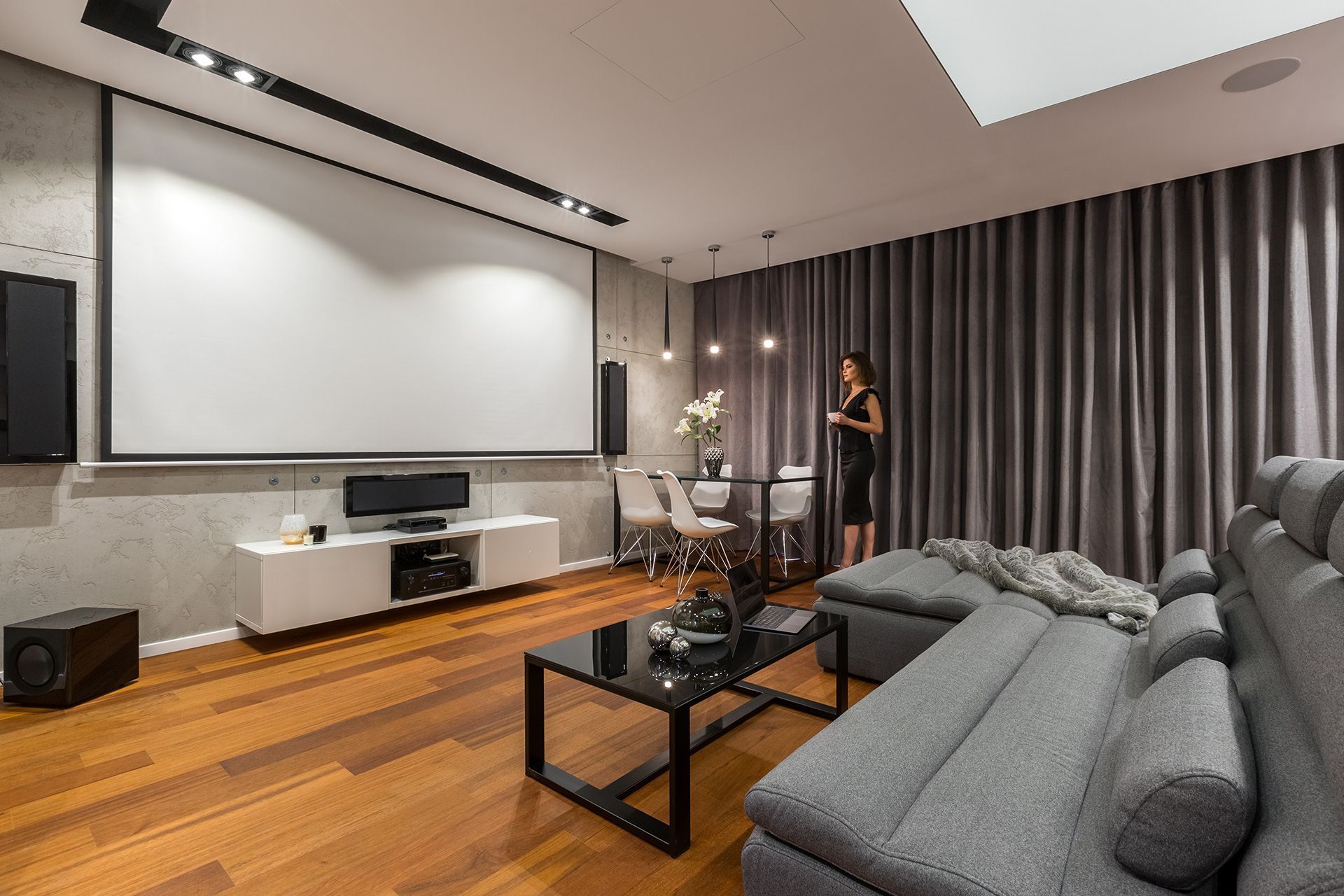 Modern living room with projector screen, grey sofa, wood floors, and a person.