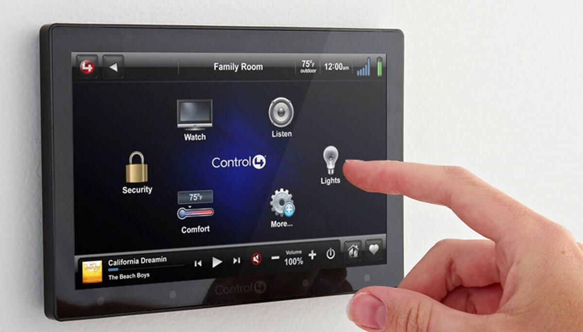 Hand touching a touchscreen control panel on a white wall, displaying smart home control icons and a music player.