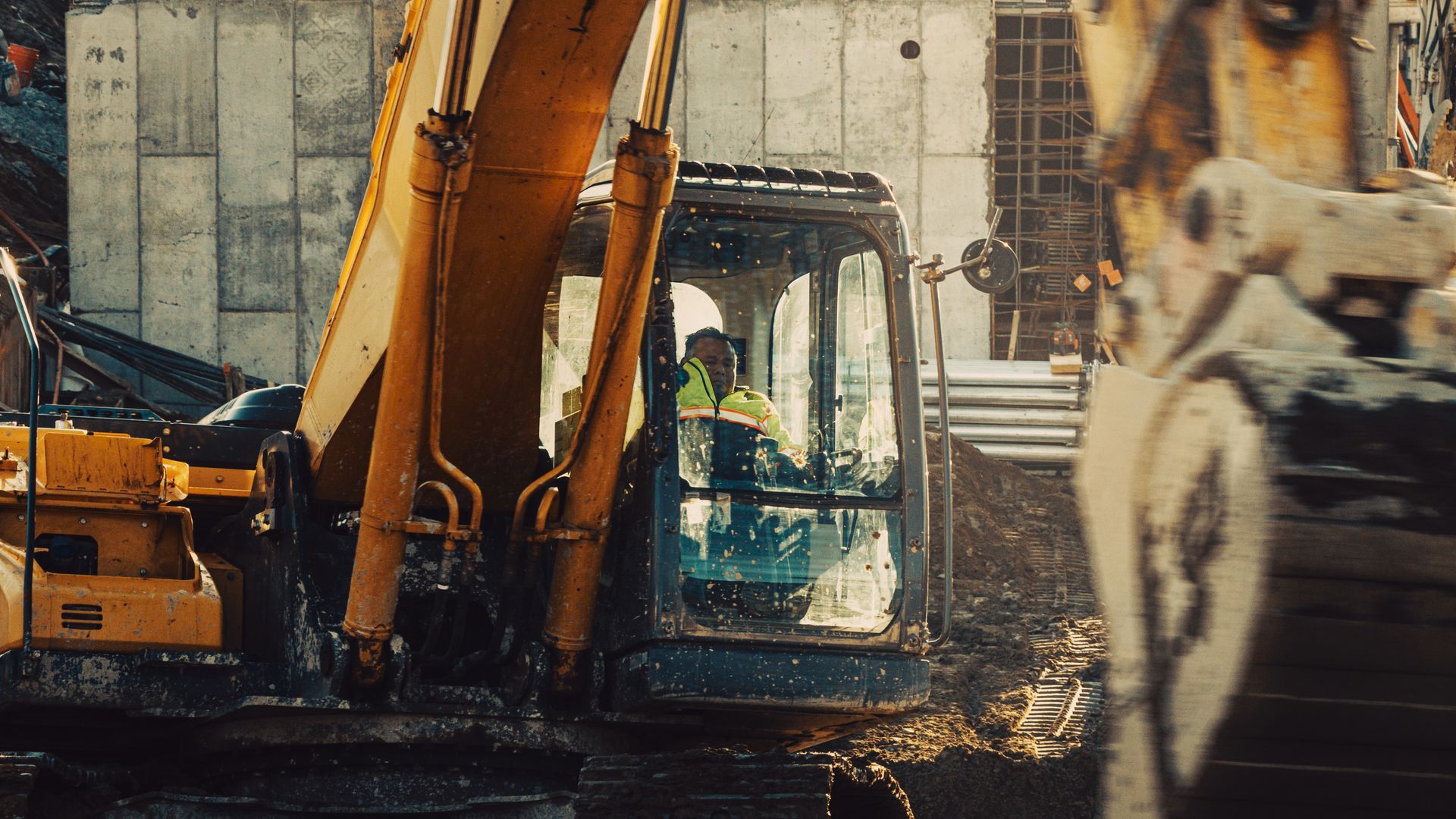 Construction worker operates an excavator on a muddy construction site; yellow machine, buildings in background.