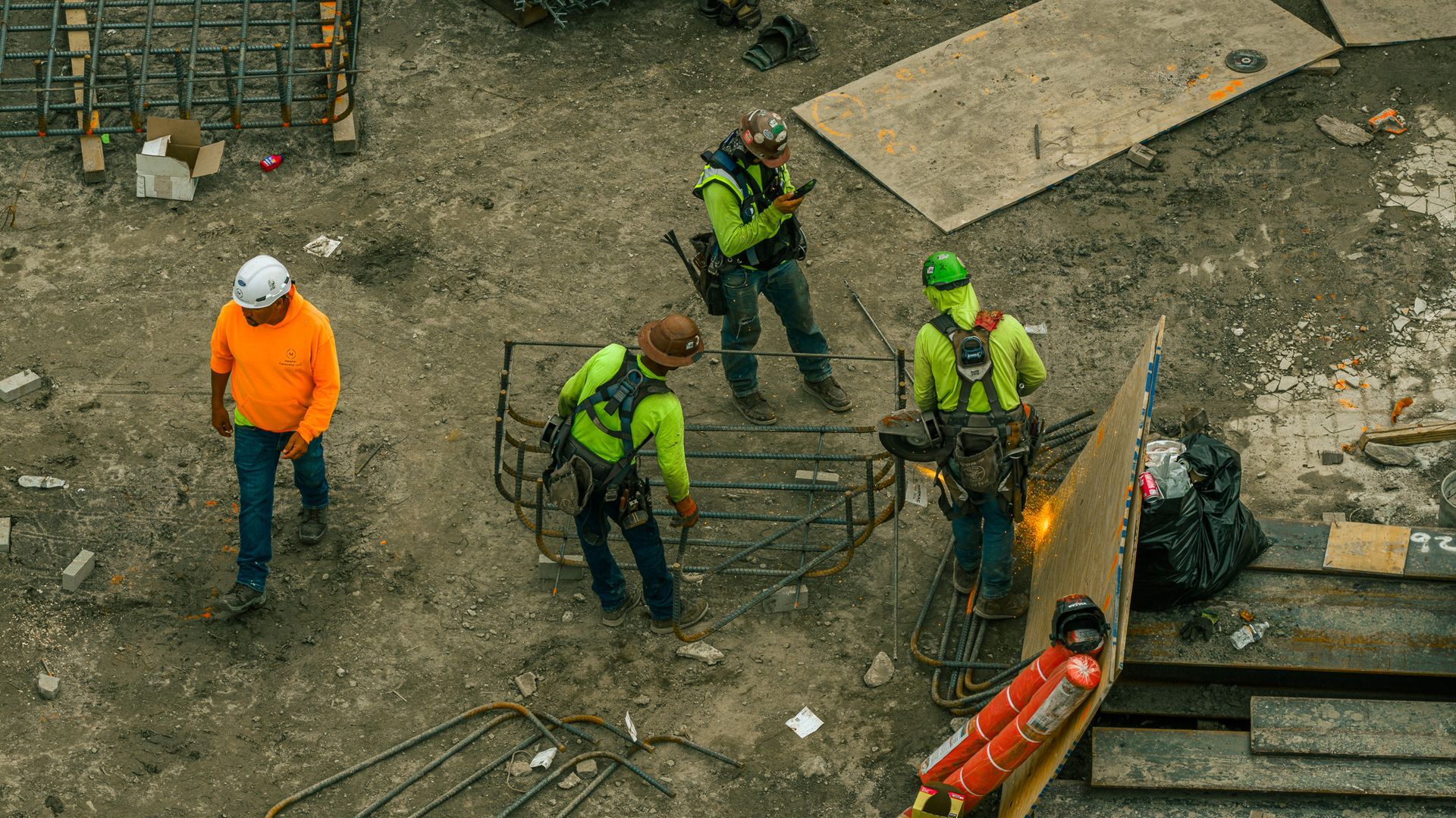 Construction workers welding on a concrete site. One in orange, three in green, near equipment and debris.
