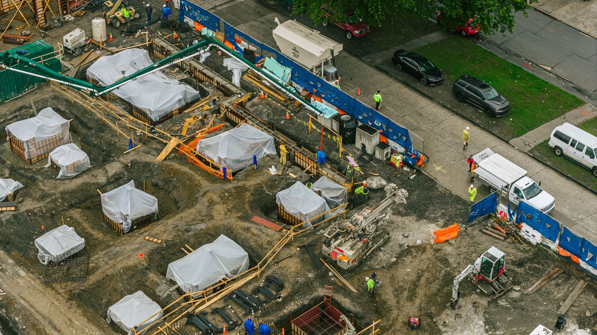 Construction site with covered structures, machinery, and workers. Blue fencing lines the perimeter near a street.