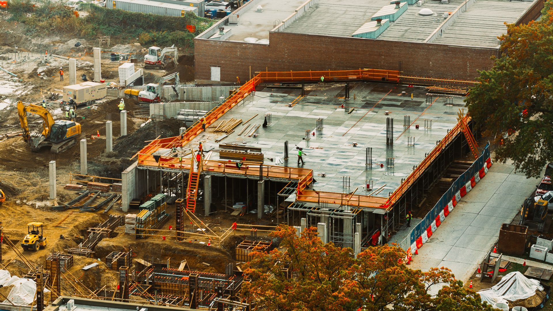 Construction site: concrete foundation and pillars, orange safety barriers, heavy machinery, workers.