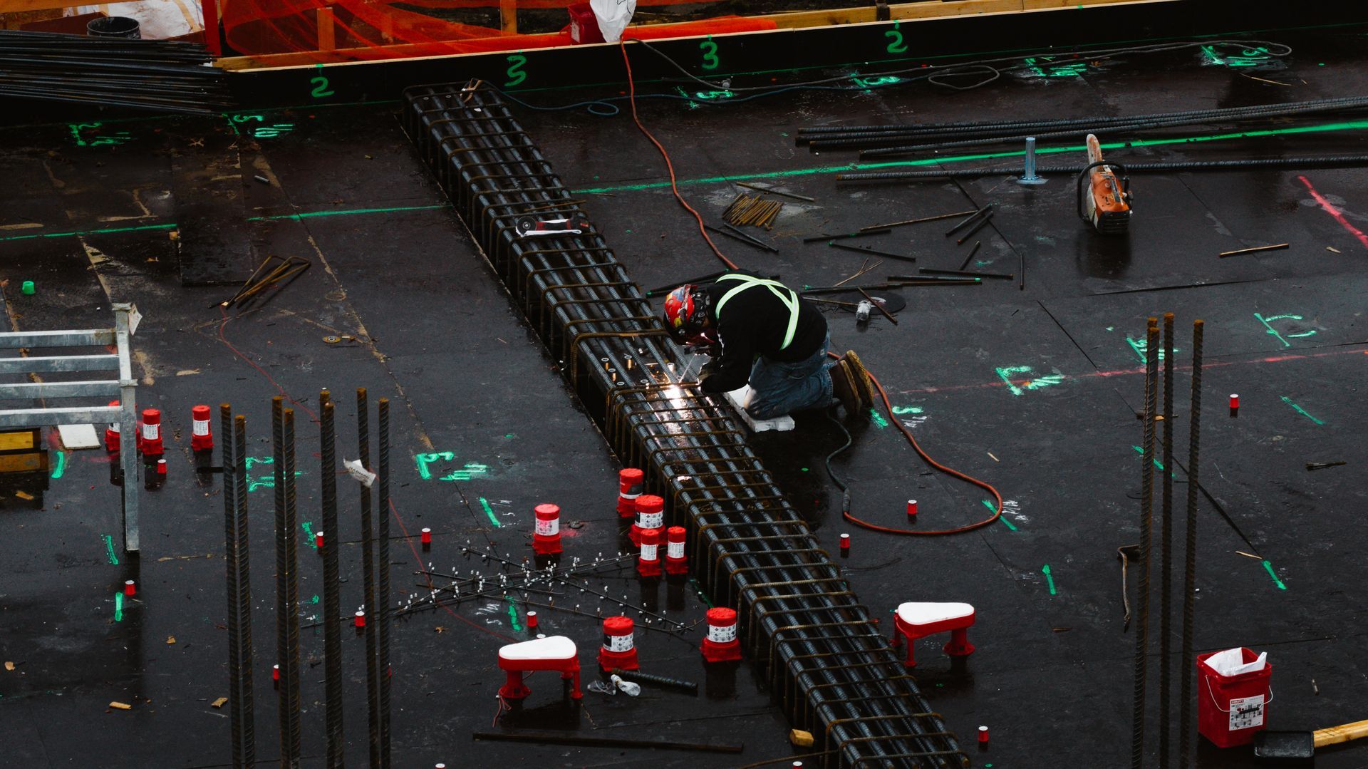 Construction worker welding steel rebar on a dark, wet surface. Sparks fly.