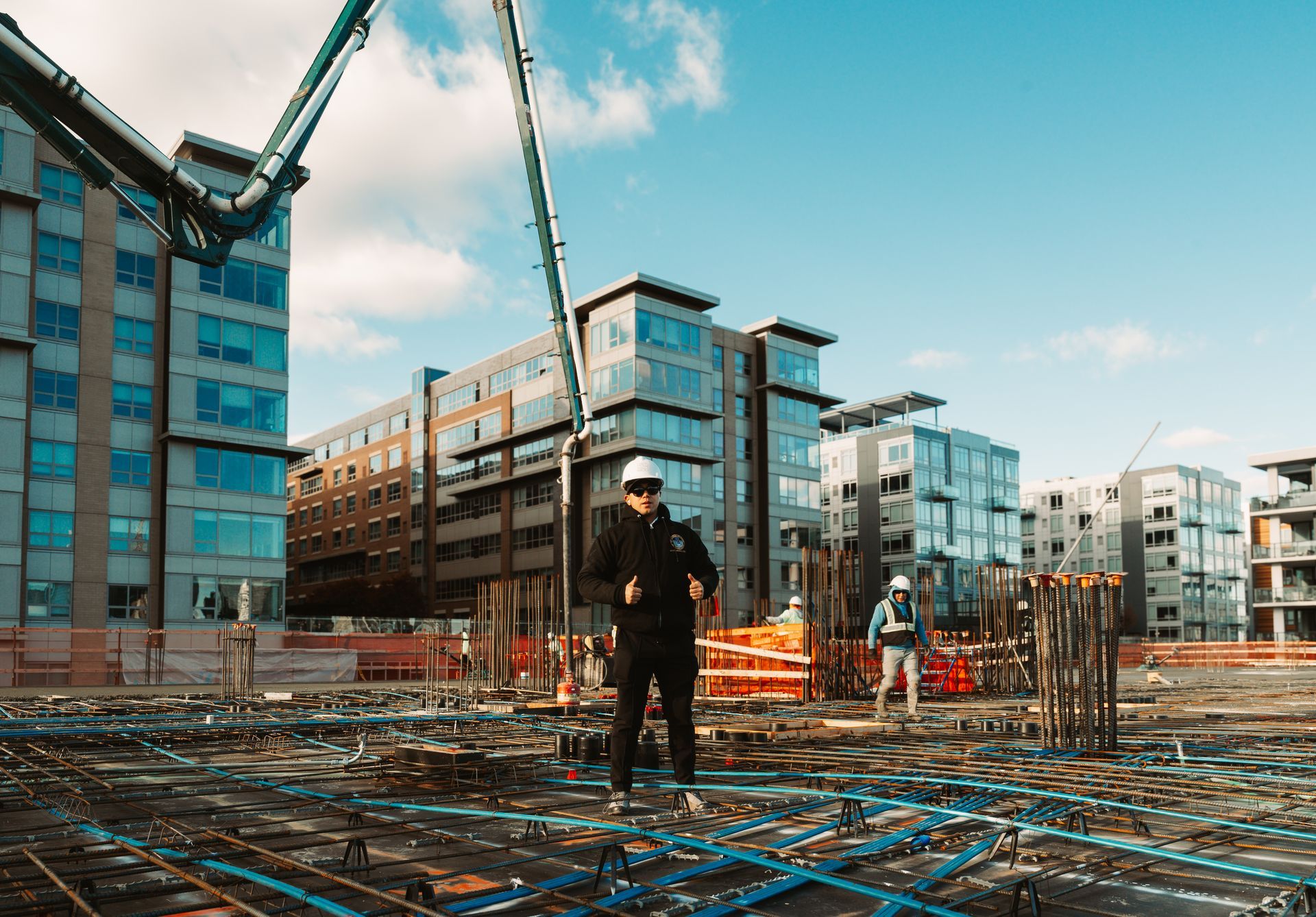 Construction workers on a concrete foundation, buildings in background, blue sky, concrete pump arm extended.