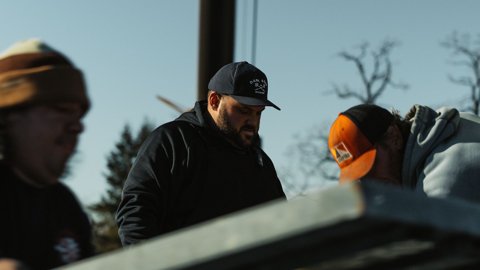 Three men working outdoors, wearing hats. One wears a navy hat, looking down. Clear, sunny day.