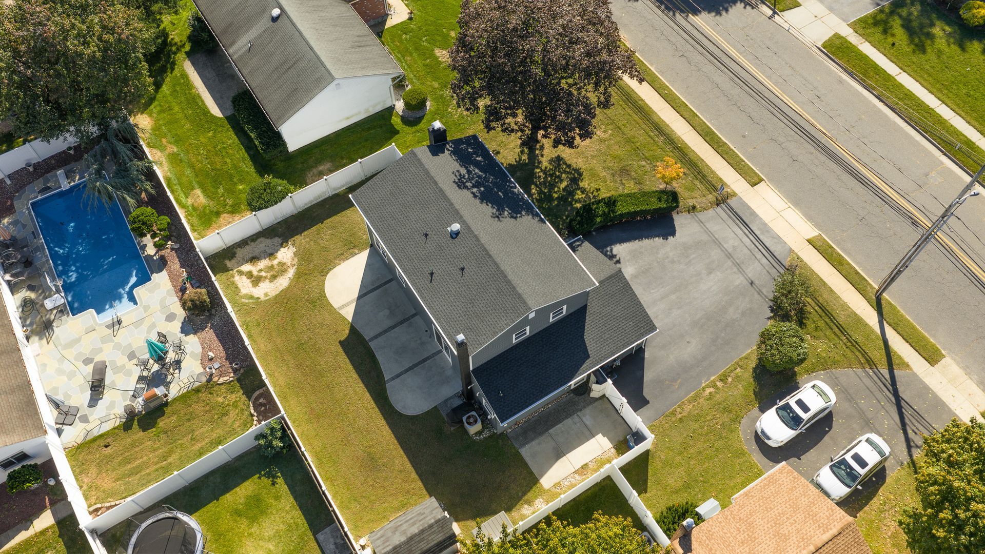 Aerial view of a house with a driveway, yard, and pool. Two cars are parked on the driveway near the street.