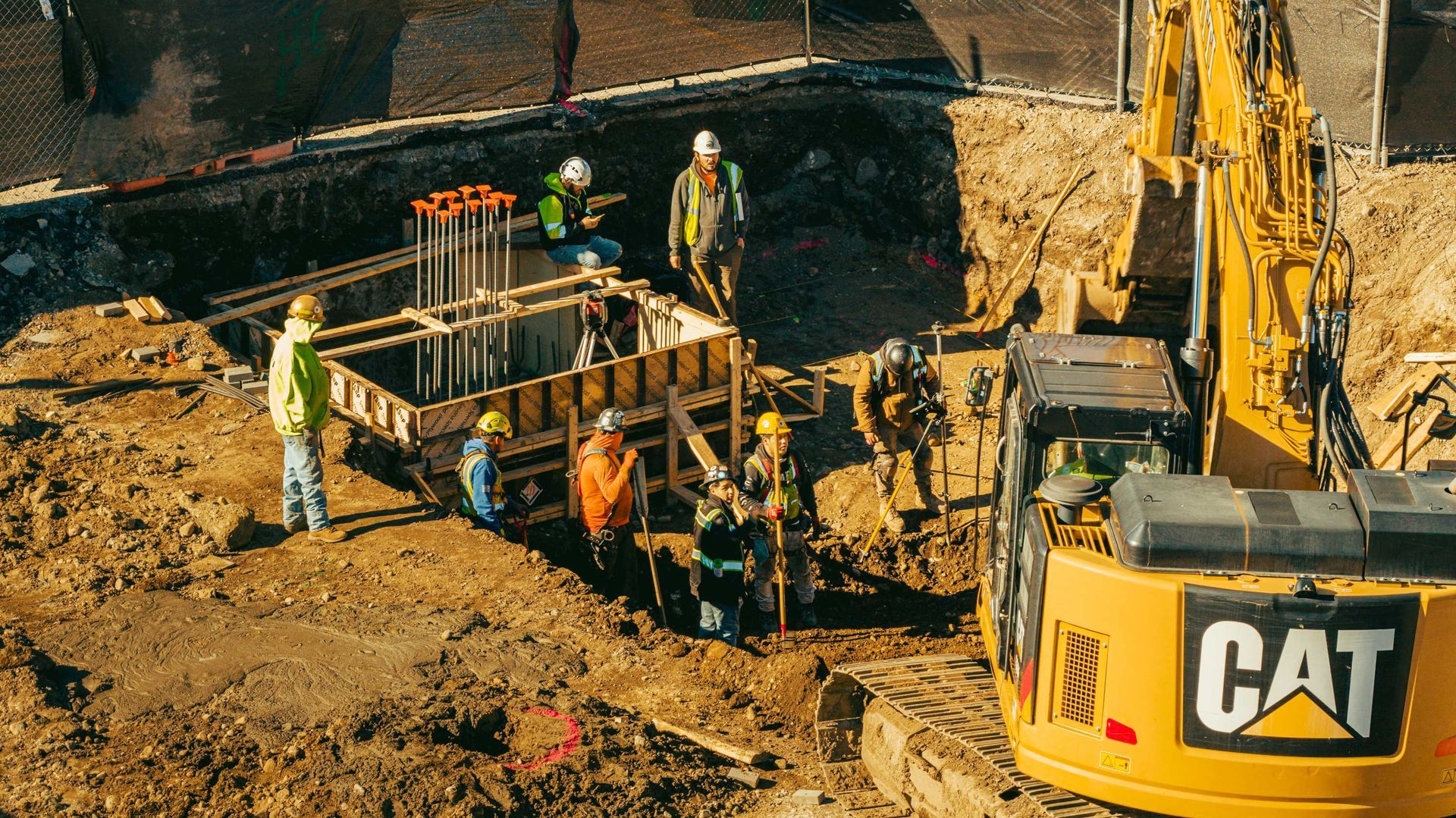 Construction site with workers around a foundation and an excavator. Soil is brown. Yellow CAT excavator.