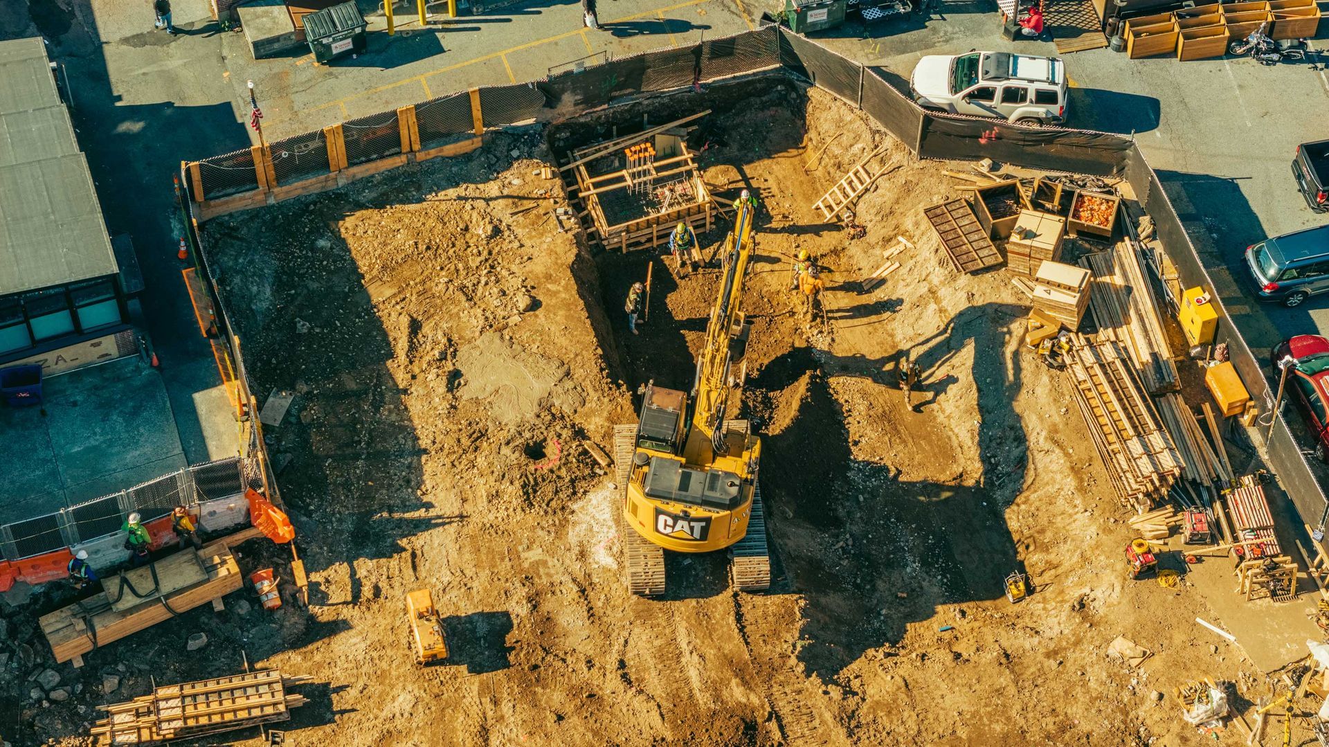 Construction site with excavator digging in a dirt pit, surrounded by a fence and materials.