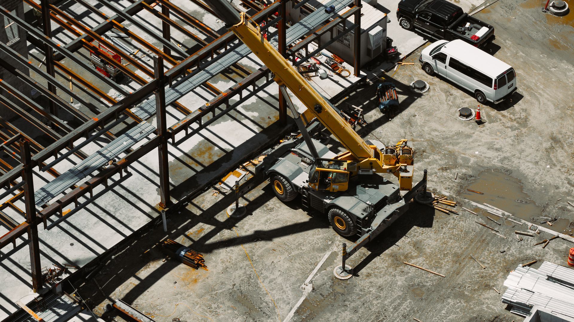 Yellow crane lifting steel beams at a construction site. Gray concrete and metal framework visible. Cars parked nearby.