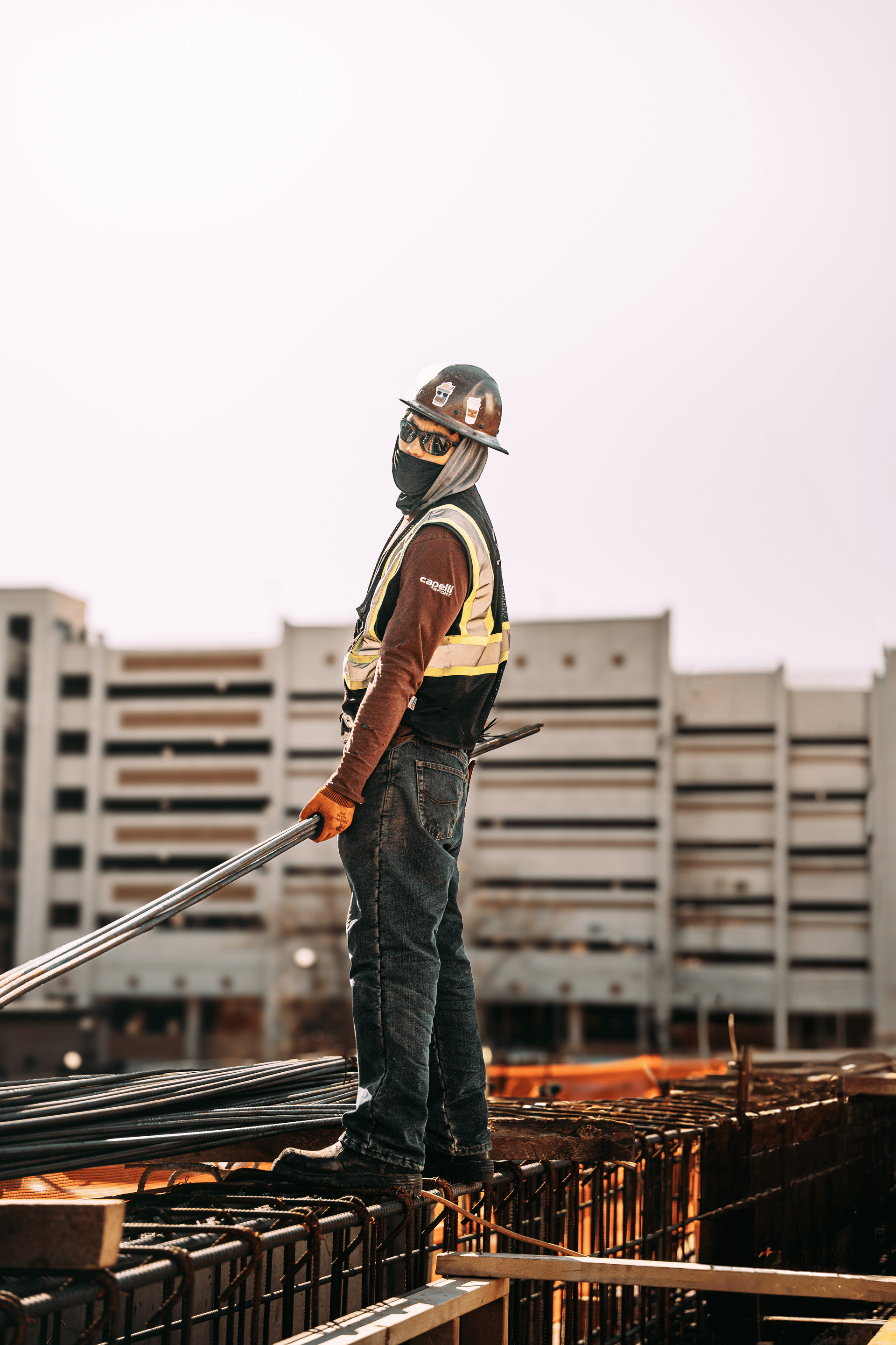Construction worker on a rooftop, holding rebar, wearing safety gear, with buildings in the background.