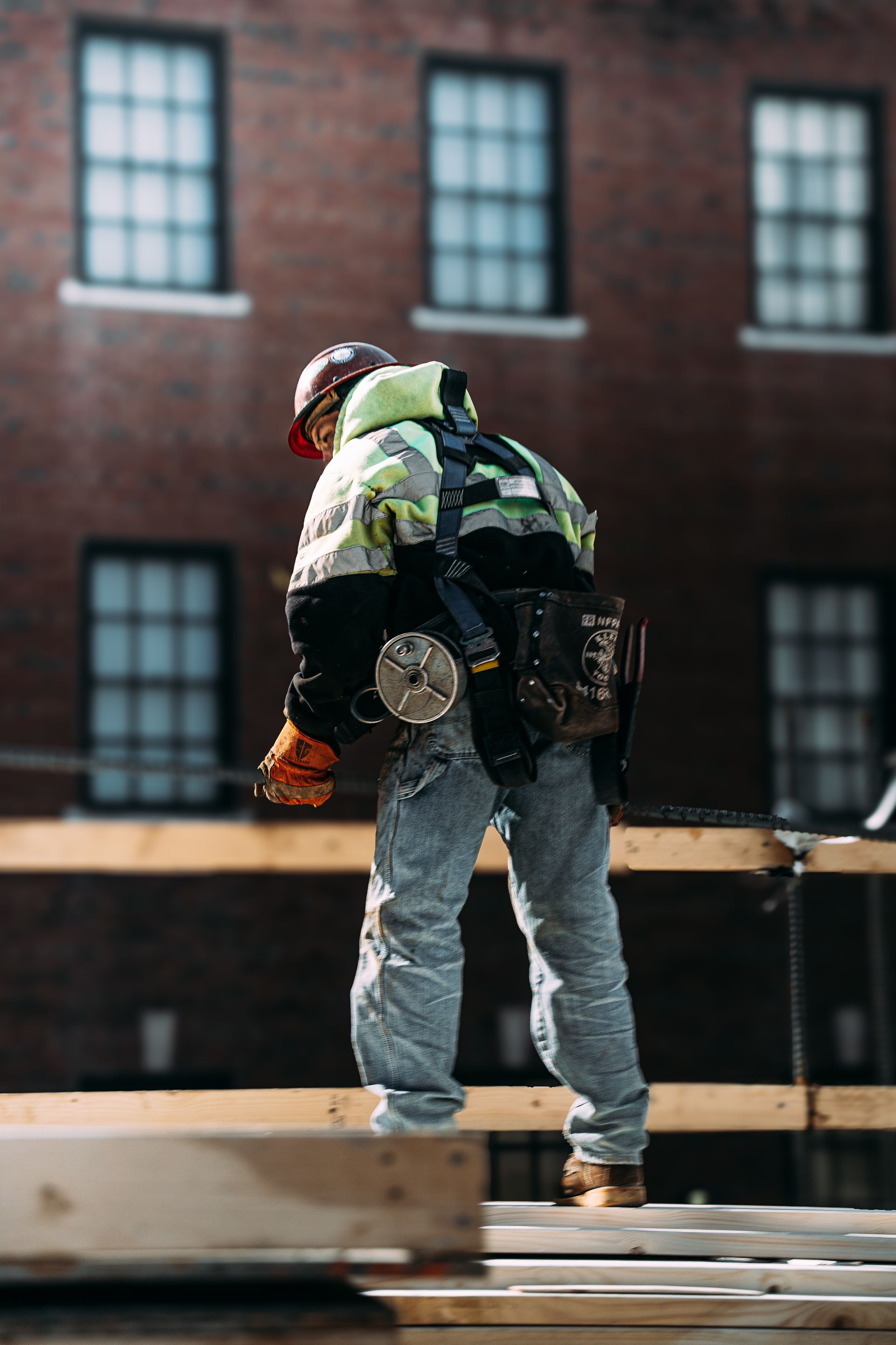 Construction worker on wooden beams, wearing safety gear, working on a brick building exterior.