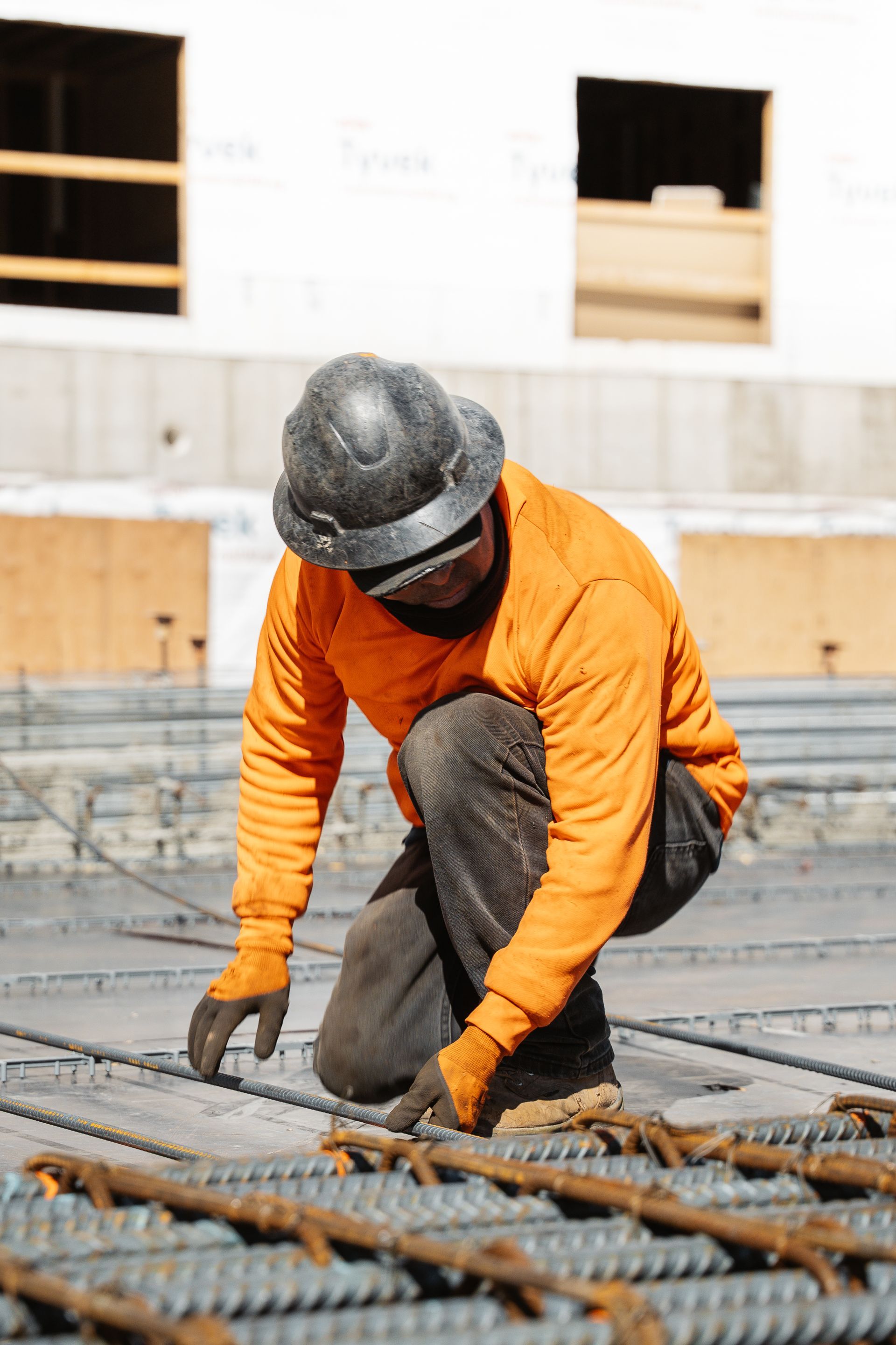 Construction worker in orange shirt and hard hat inspects rebar on a building site.