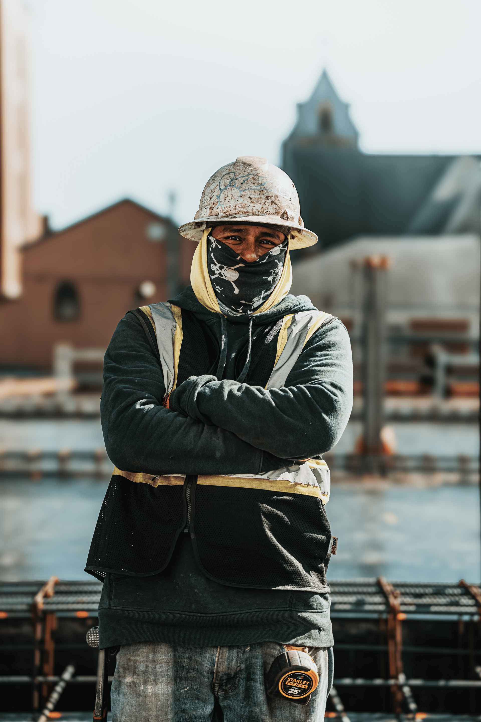 Construction worker with arms crossed, wearing a hard hat, reflective vest, and face covering, outdoors.