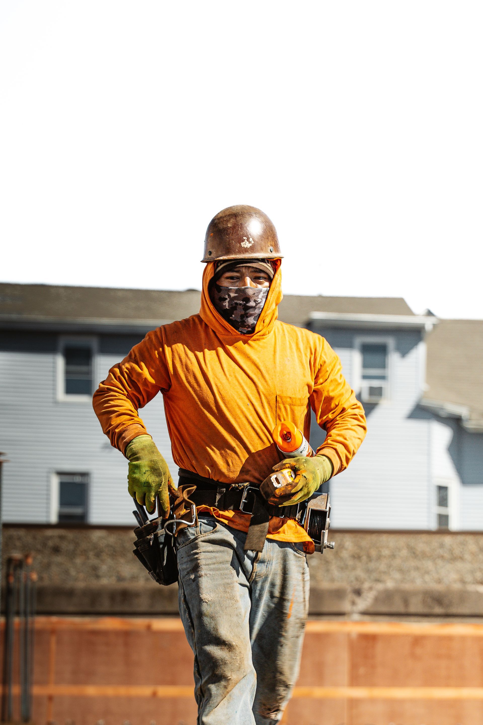 Construction worker wearing hard hat and orange hoodie on rooftop, hands on hips, face smudged.