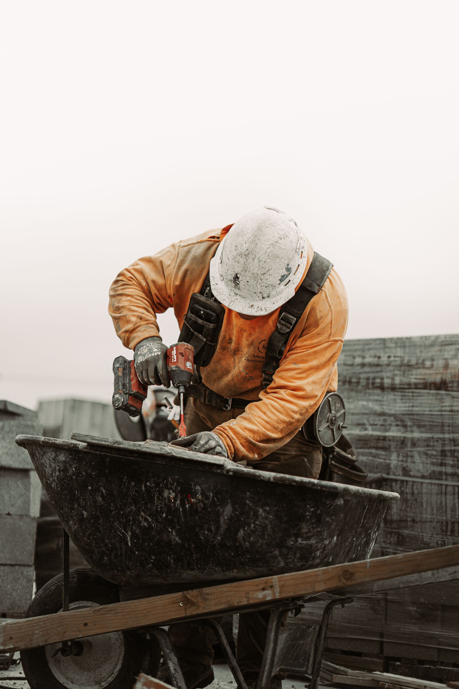 Construction worker drills into a wheelbarrow, wearing an orange jumpsuit and hard hat.