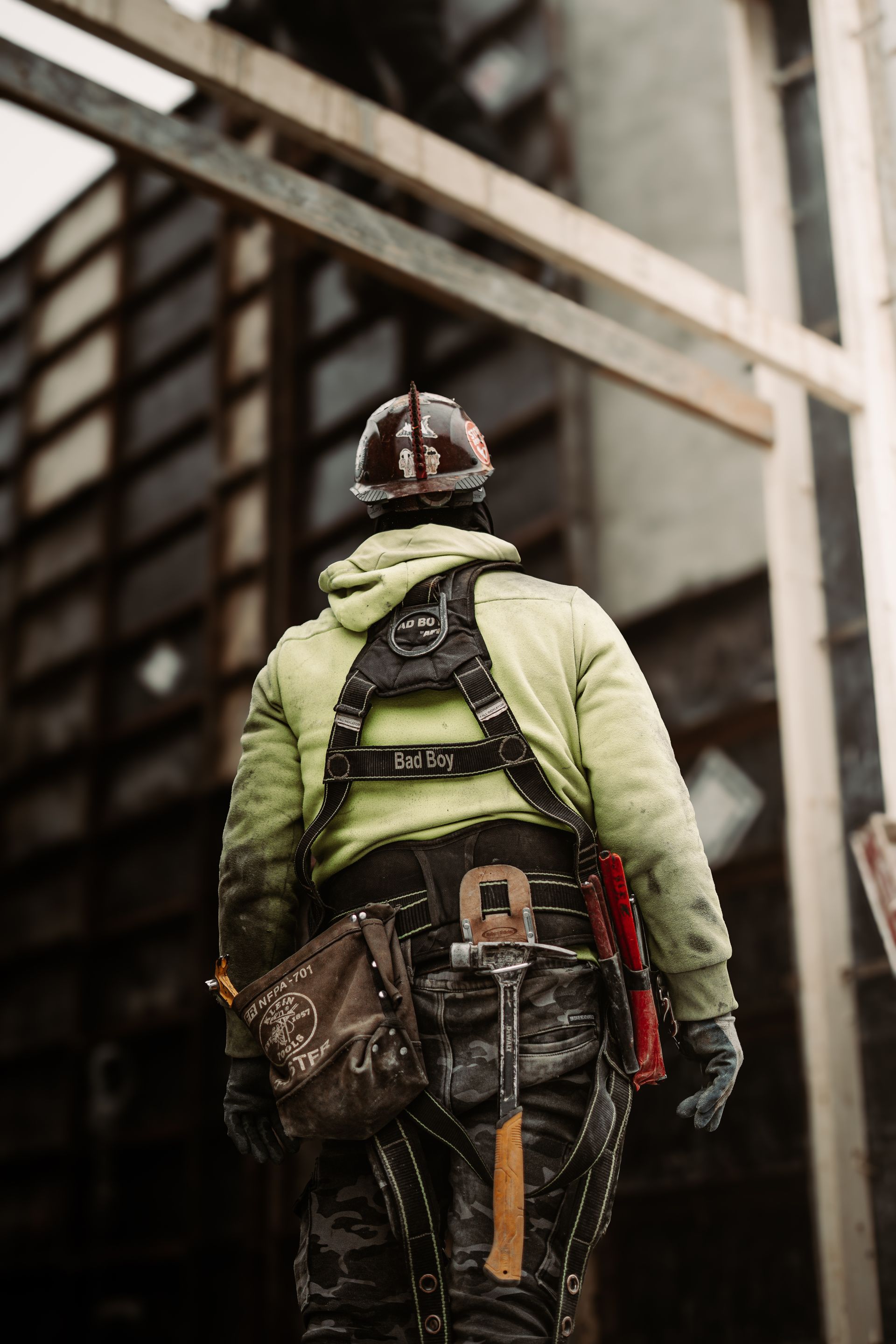 Construction worker in green jacket, hard hat, and harness, walking toward a building under construction.