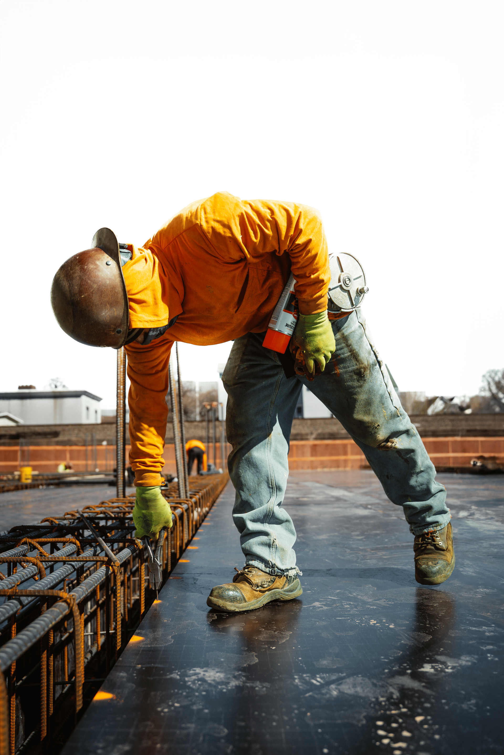 Construction worker inspecting rebar on a construction site. Wearing orange jacket, hard hat, and gloves. Leaning over.
