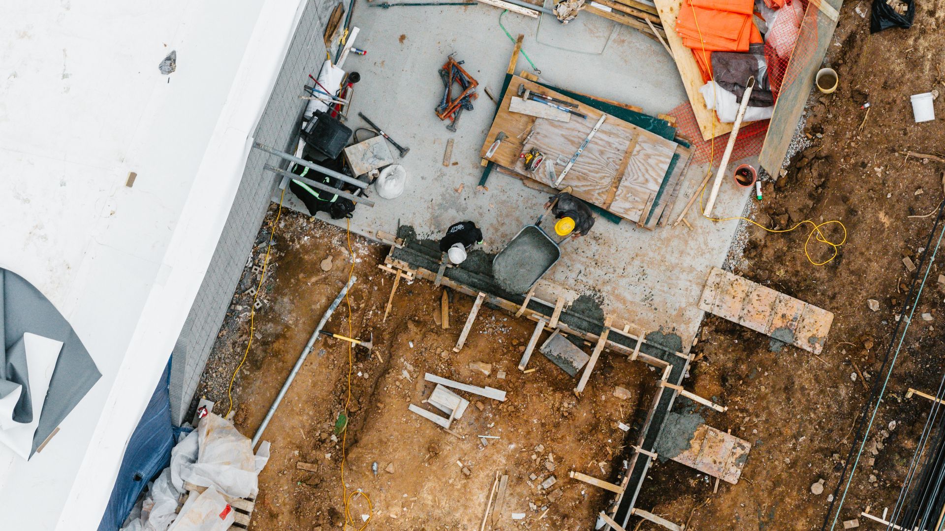Construction site aerial view; workers near building foundation. Dirt, wood, tools, and materials are visible.