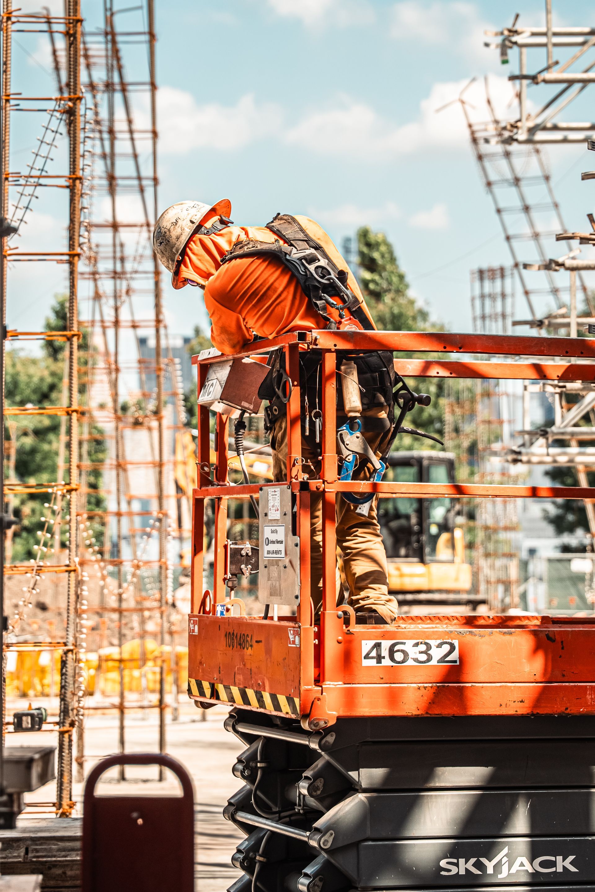 Construction worker in an orange lift platform near metal scaffolding, inspecting something.