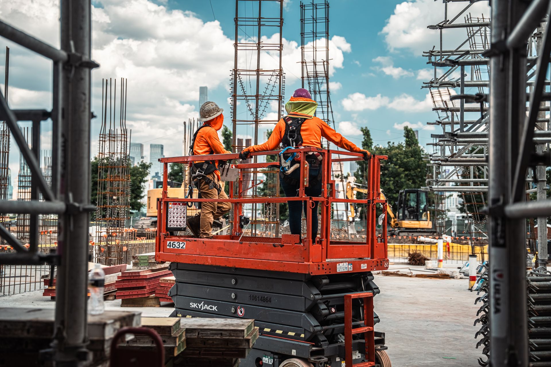 Two construction workers on an orange lift. They are wearing orange vests and hard hats, amidst a construction site.