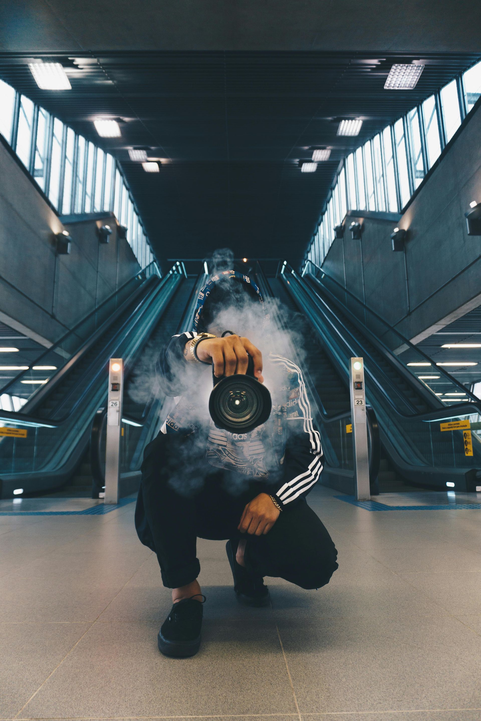 Person holding a camera, engulfed in smoke, squats in front of escalators in a subway station.