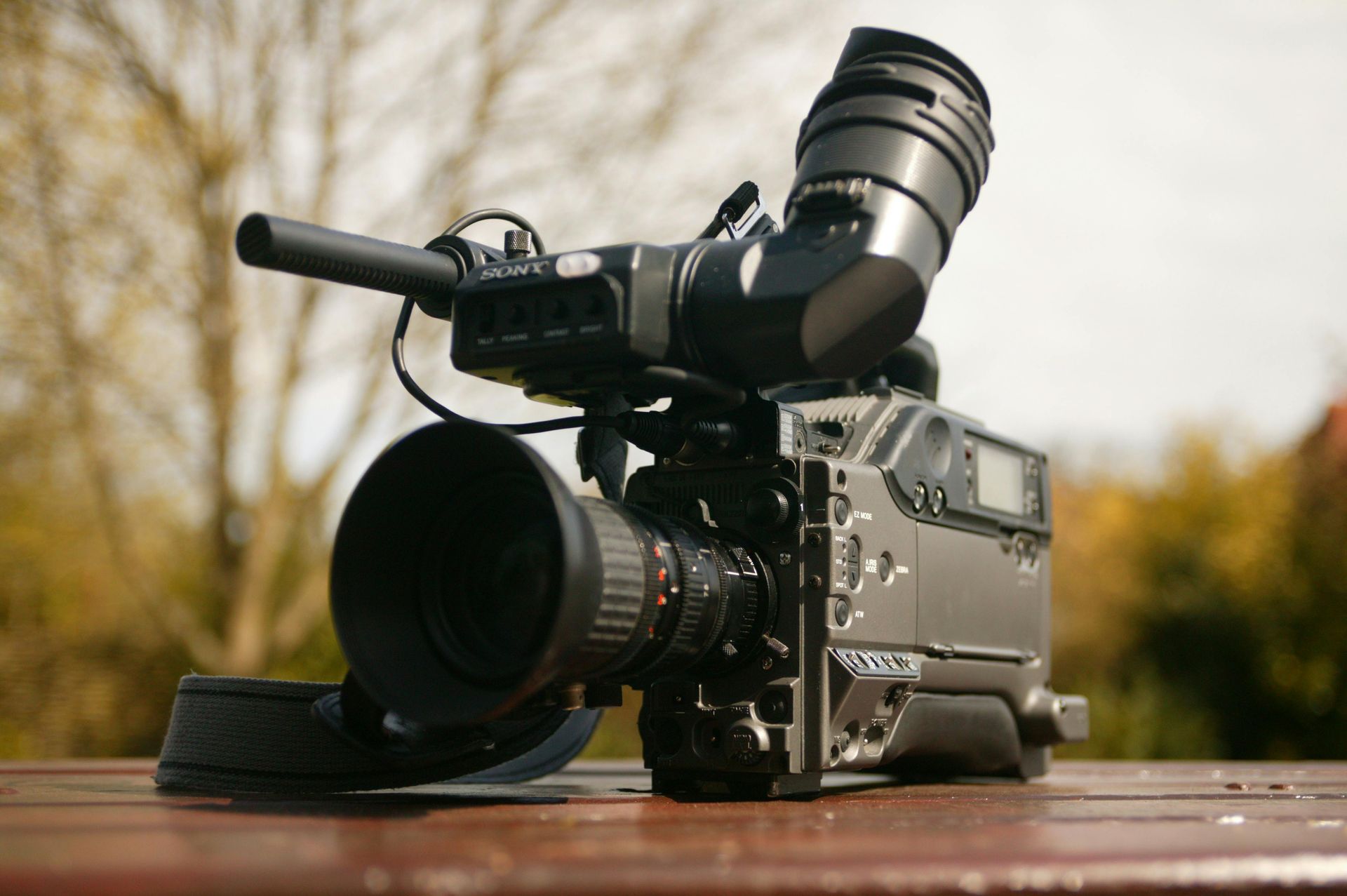 Black professional video camera with large lens sitting on a wooden table, outside with blurred background.
