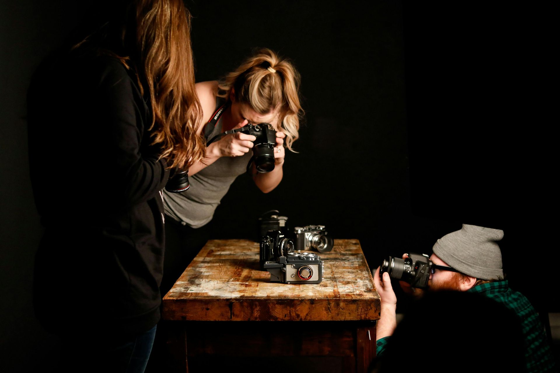Three people photographing vintage cameras on a wooden table in a dimly lit room.