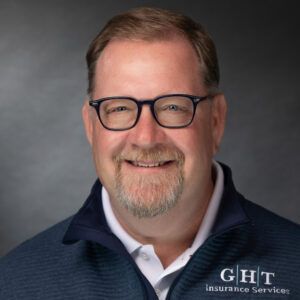 A man with glasses and a beard is wearing a jacket that says ght insurance services