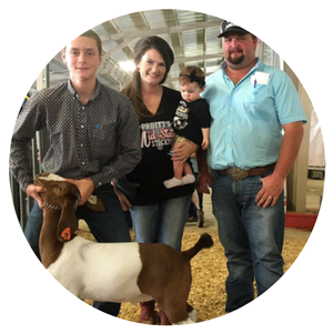 A family stands in an indoor arena with a goat. A young person holds the goat's head while an adult holds a small child.